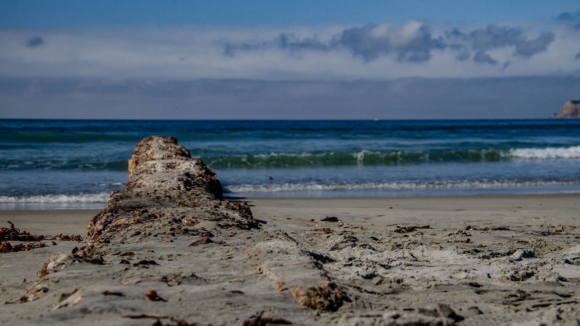 The image shows a long, weathered, thin concrete and stone jetty on a sandy beach. The jetty extends out from the lower left corner of the image and continues into the distance. It is covered in sand and seaweed and has a rough, textured surface. The beach is wide and sandy, with waves gently lapping at the shore in the background. The ocean is a clear blue, with whitecaps on the waves. The sky is a light blue, with a few fluffy white clouds. The overall feel of the image is peaceful and serene.  A small, rocky cliff is visible in the very distant background, behind the ocean.