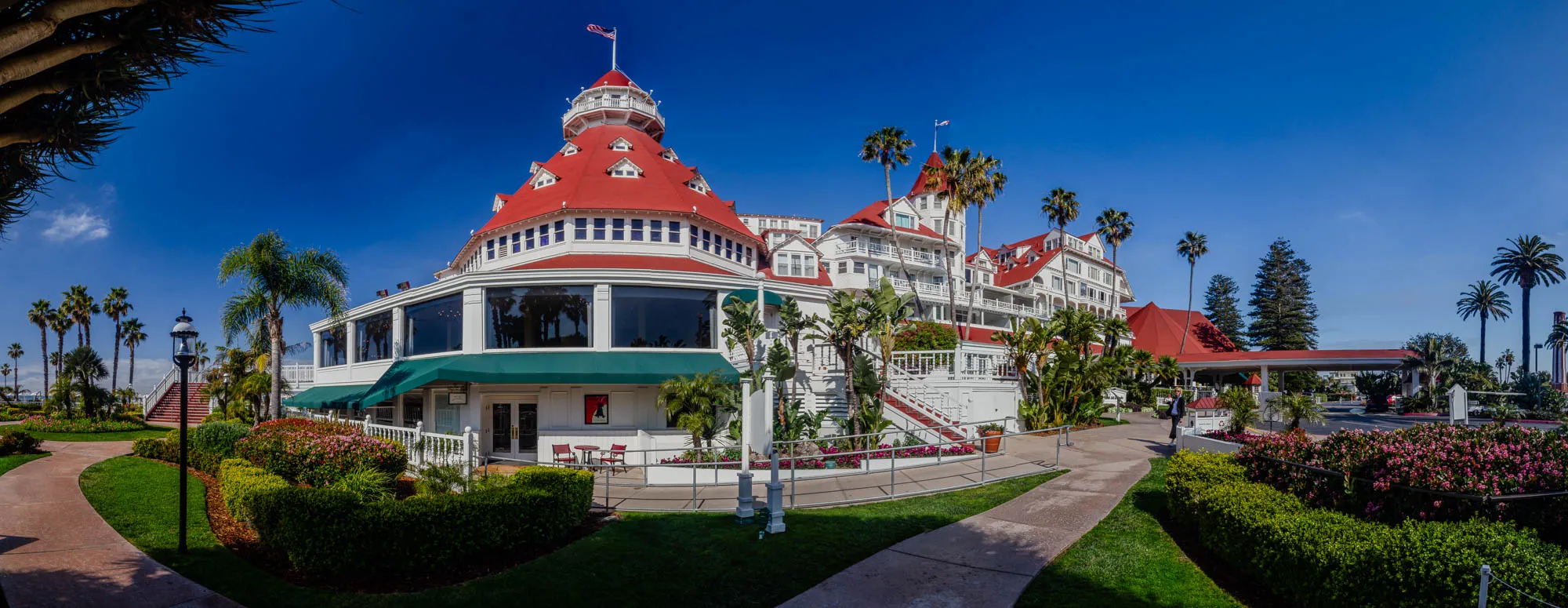 The image shows a large, white, red-roofed hotel building with an American flag on top. It is a multi-story building with many windows and balconies. The building is surrounded by lush green landscaping, including palm trees, bushes, and grass. In front of the building is a paved walkway, and there is a lamppost on the left side of the image.  The building appears to be in a resort setting. There is a wide, grassy lawn in front of the building, with a winding pathway leading away from the viewer.