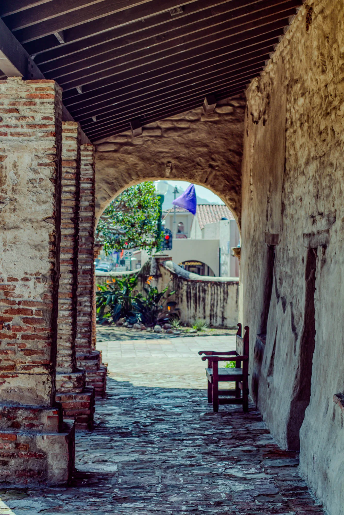 The image shows a brick walkway leading through a stone archway. On the left side of the walkway are a series of brick pillars.  On the right side of the walkway is a rough stone wall.  There is a wooden bench sitting against the wall to the right.  The archway leads to a courtyard, and a purple flag can be seen fluttering in the breeze on a flagpole in the distance.  There is some foliage, a white building, and a red traffic light visible in the distance. The walkway is made of flat, irregularly-shaped stones.  The archway is made of a darker stone, and the pillars have a rough, uneven texture.