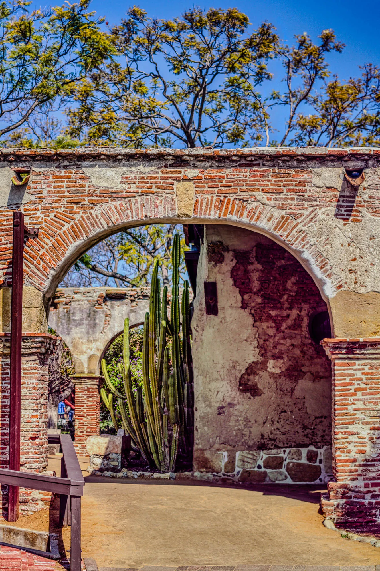 The image shows an arched brick doorway with a tall cactus plant growing in the middle of the passage.  The archway is made of red brick and the wall behind it is a light brown color with some patches of plaster missing to reveal the red brick underneath.  There is a second archway behind the main archway which is partially obscured by the cactus.  The background of the image is a blue sky with a few branches of a tree visible at the top of the frame.  In the foreground, there is a brown wooden handrail and a small part of a brick pathway is visible.  A person is visible walking through the passage, but their face is obscured.