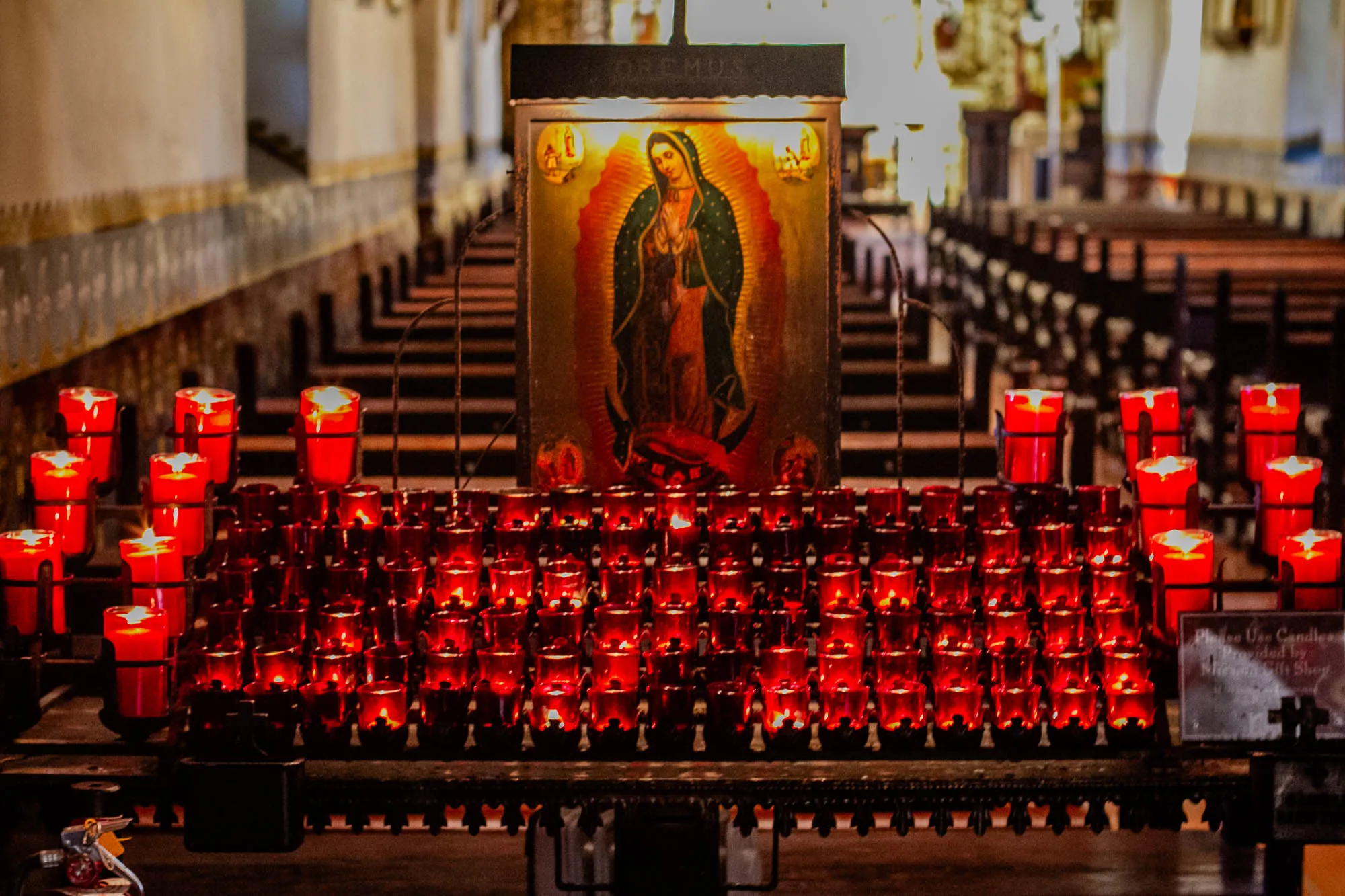 The image depicts a church interior. There is a large, framed painting of a religious figure, a woman wearing a blue robe and holding her hands in prayer. The painting is illuminated by a light source from above.  In front of the painting, a row of about 50 red votive candles are lit and sitting on a metal candle holder, their flames casting a warm glow on the surrounding area. The votive candles are arranged in a rectangular shape, creating a visual focal point in the image.  The church interior appears to be spacious and dimly lit, with wooden pews visible in the background. The image exudes a sense of reverence and spirituality.