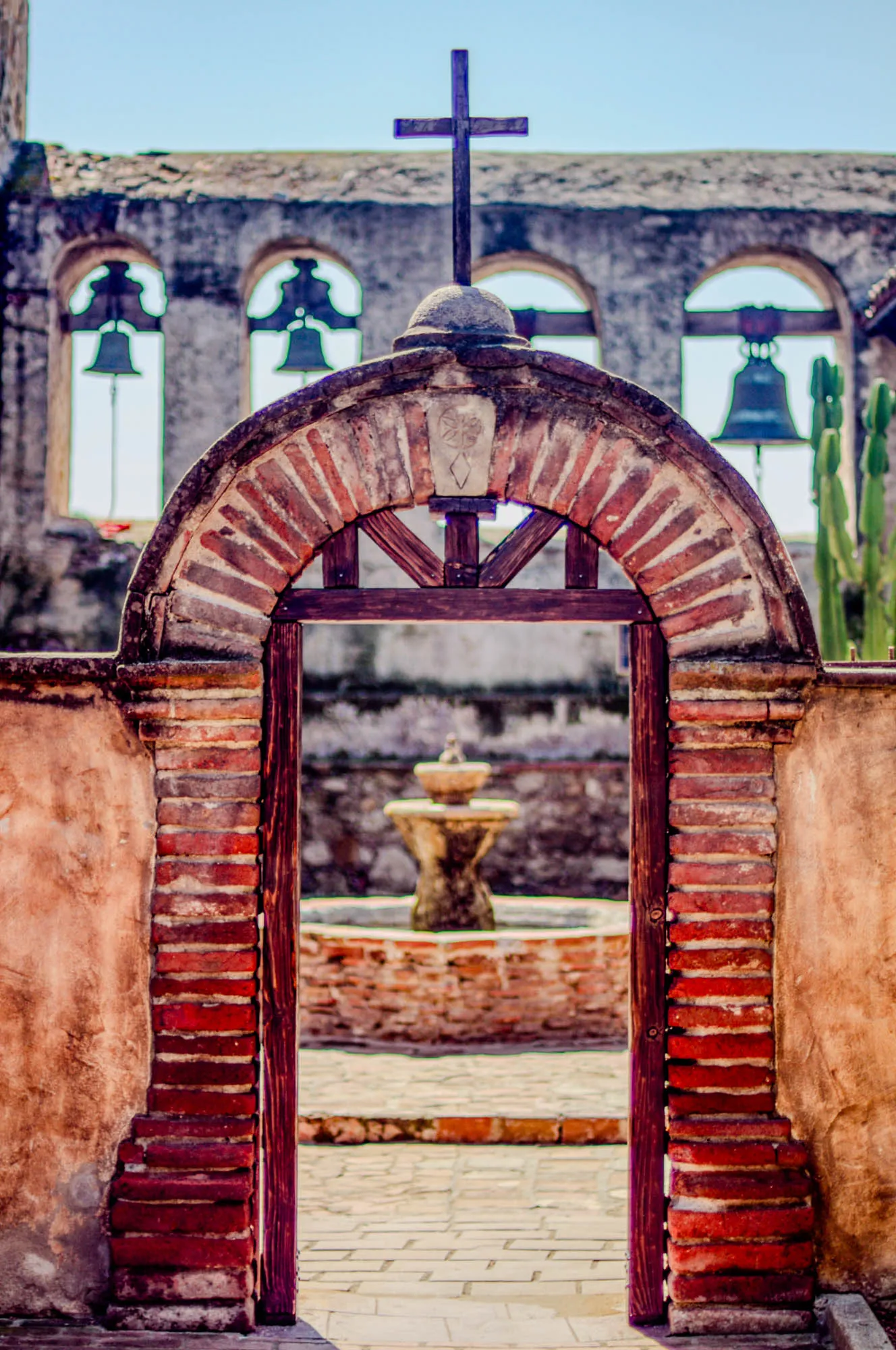 The image shows a brick archway with a wooden door leading to a courtyard. The archway is made of red bricks and has a wooden cross on top. The door is made of dark wood and has a simple design. There are three bells hanging in the windows above the archway. There is a fountain in the center of the courtyard. The background is a wall with a stucco finish.  The image is taken from a low angle and has a warm, sunny tone.