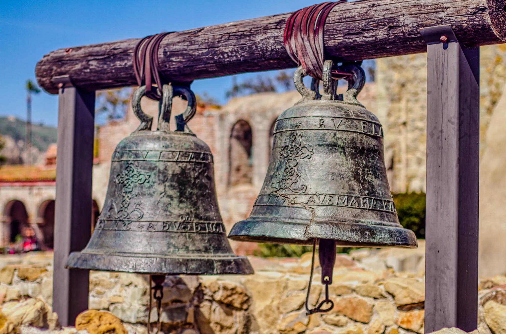 The image shows two large, weathered bronze bells hanging from a wooden beam. The bells are attached to the beam by thick brown leather straps. The bells are hanging from a metal frame that appears to be bolted to the wooden beam. The bells are facing the viewer and the frame is on the right side of the image. The bells appear to have a faded patina.  They are both inscribed with lettering. The bell on the left has an inscription that is partially hidden from view by the beam. The inscription on the right bell is visible and reads "R.V.E.M." and "1716.AVE MARIA" in faded lettering.  Behind the bells, there is a stone wall and trees. The stone wall appears to be an old building, and there is a faint figure standing in the doorway. The background is slightly blurred.