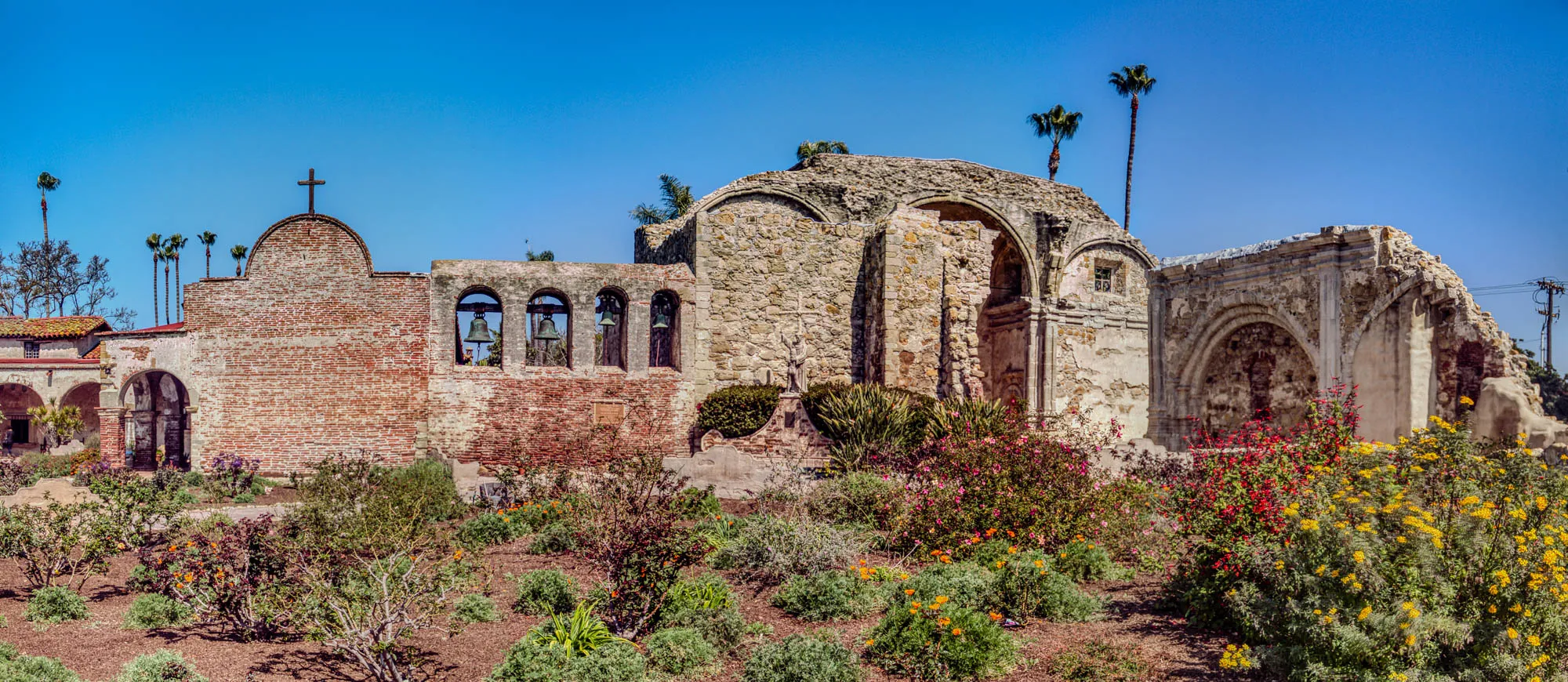 The image shows a weathered, stone and brick building. A portion of the building has collapsed, leaving a large, arched opening. There are several bells hanging in the building, and a small cross is visible at the top. The building is surrounded by a garden with green bushes and flowers. The sky is clear blue with some palm trees in the background. The building is likely an old church or mission.