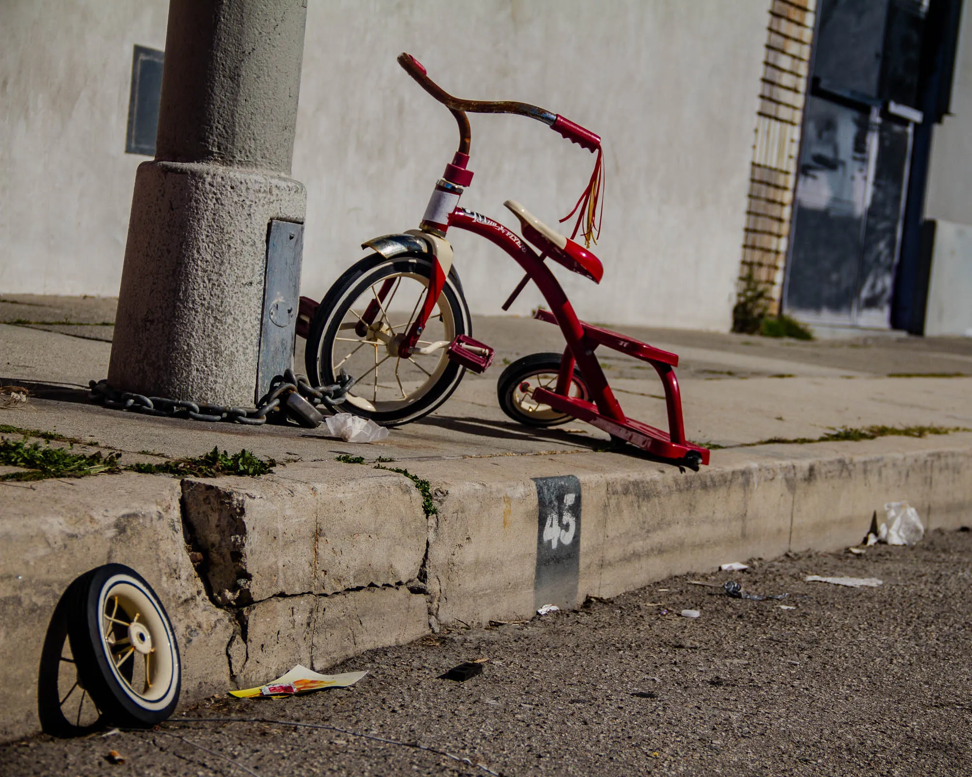 A red tricycle is chained to a concrete pillar on the edge of a sidewalk. The tricycle has a white rim with spokes on the front wheel and is leaning toward the sidewalk. The back right tire is black and the wheel is askew, the back left wheel is missing, and the seat is white and red. A chain is attached to the pillar and the tricycle, and a padlock is in the chain. The sidewalk is made of concrete and is cracked. The number "45" is painted on the curb. There are pieces of trash and the trike's missing back left wheel on the ground next to the tricycle. The background is a white wall with a black door.