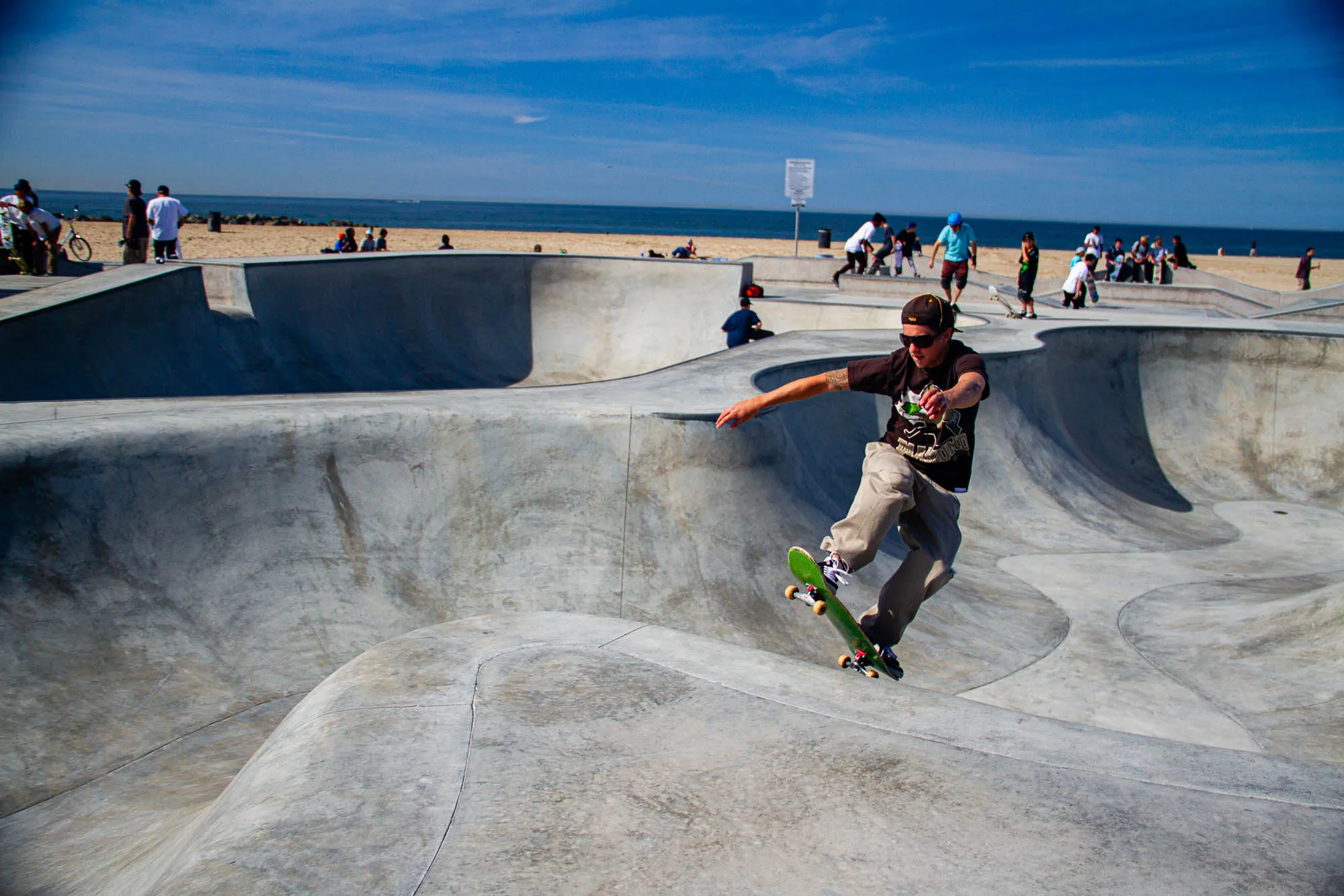 A man wearing a brown t-shirt, khaki pants, and sunglasses is skateboarding down a half-pipe in a skate park. He has one foot on the skateboard and is leaning forward.  The skateboard is green and has black wheels.  In the background are other people skateboarding and watching. The sky is blue and sunny with a few white clouds.  The beach is visible in the distance.