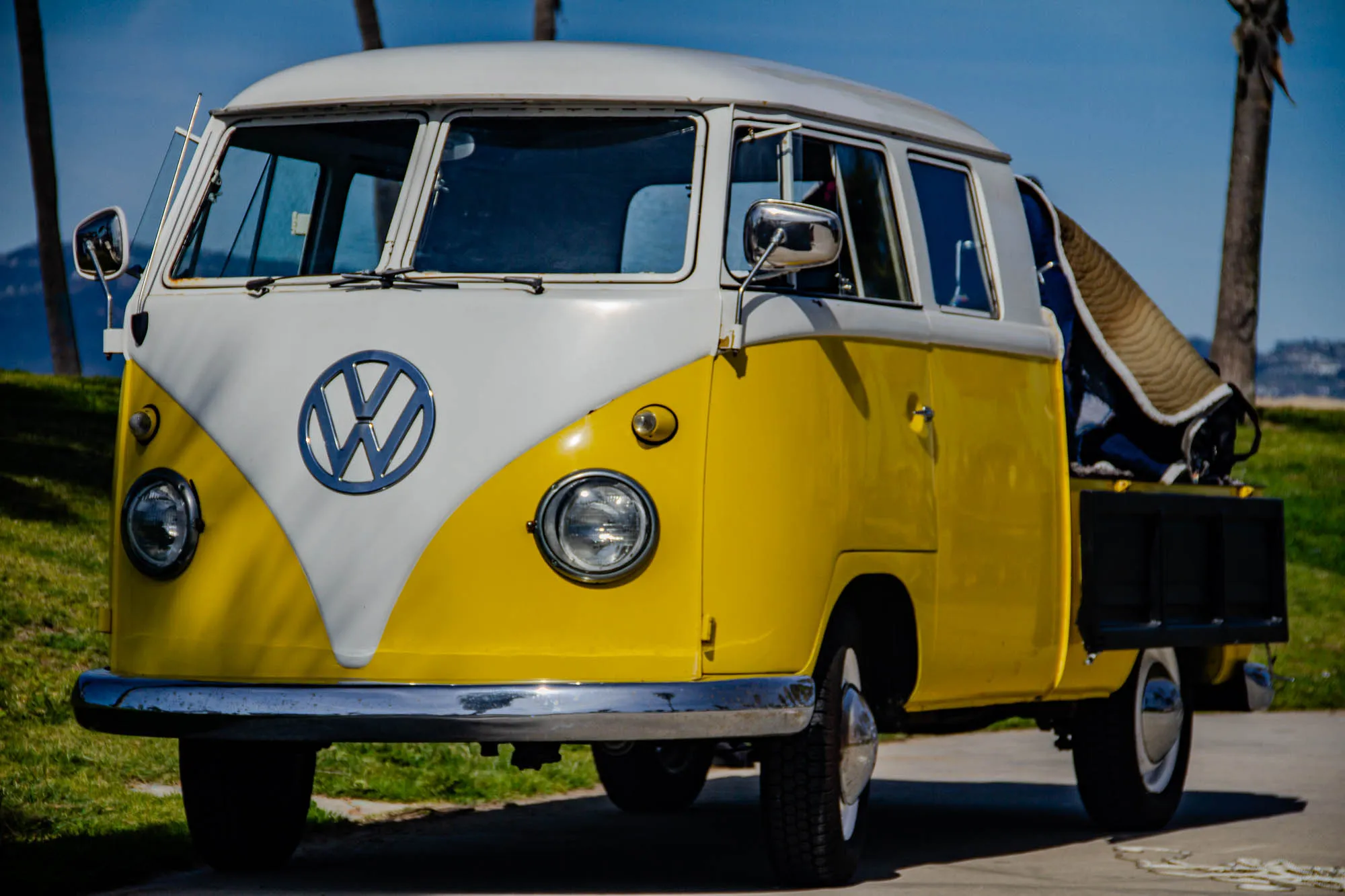 The image is of a yellow and white vintage Volkswagen van. It is a pickup truck, with a flatbed in the back. There is a large silver Volkswagen logo on the front of the van. The van is parked on a paved surface with green grass in the background. It is a sunny day, and the sky is blue. The van has a large rearview mirror on the driver's side. A blanket or tarp is draped over the bed of the truck. The van has a large windshield and side windows. There are two palm trees visible in the background.  