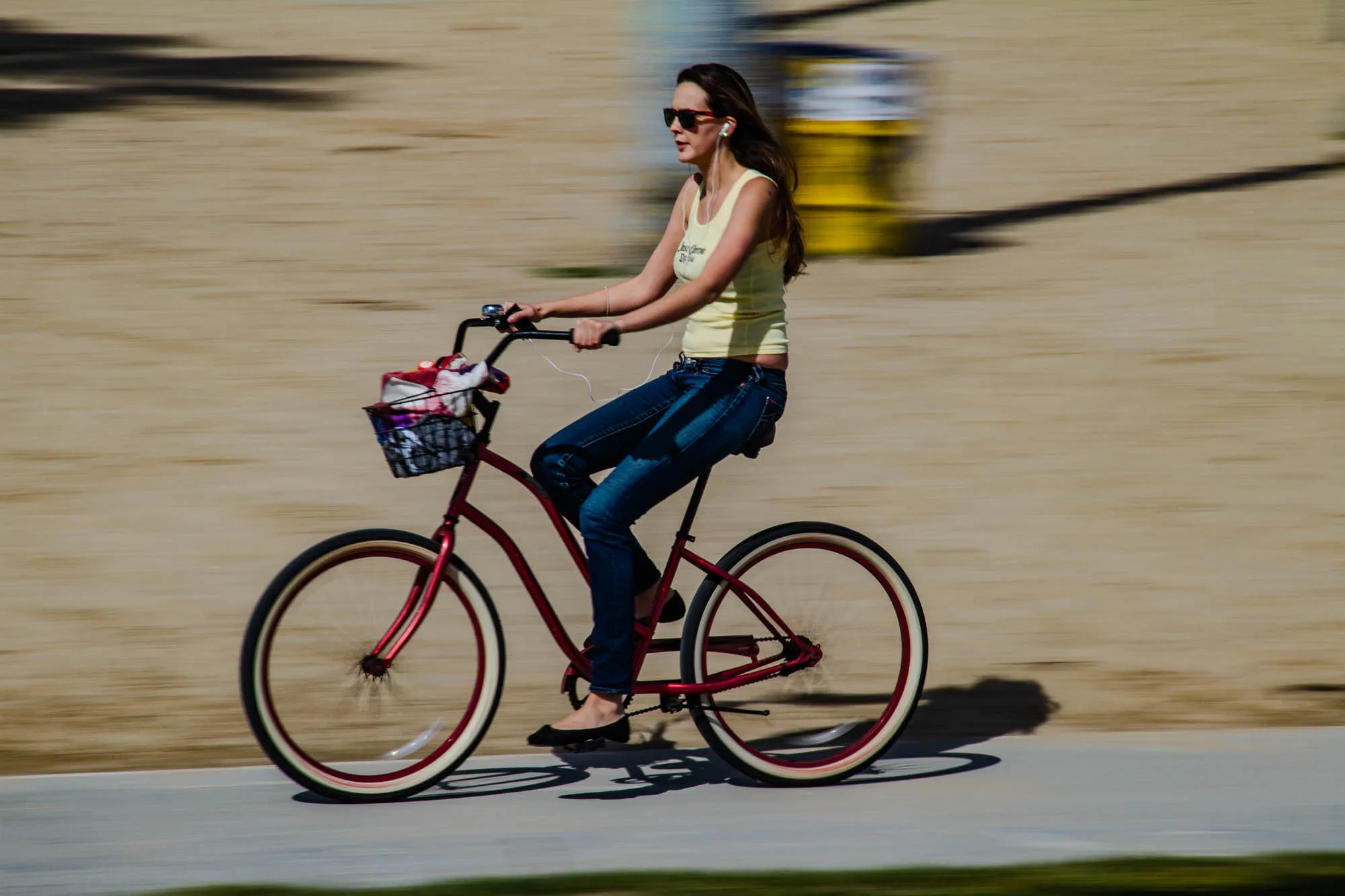 The image shows a woman riding a bicycle on a paved path. The woman is wearing blue jeans and a yellow tank top. She has dark hair and is wearing sunglasses. She is riding a red bicycle with a white rim on the wheels. There is a basket on the front of the bicycle with a pink and white fabric inside. The path is made of concrete and there is grass on both sides of the path. The woman is moving towards the right side of the image, so her image is slightly blurred as if in motion.