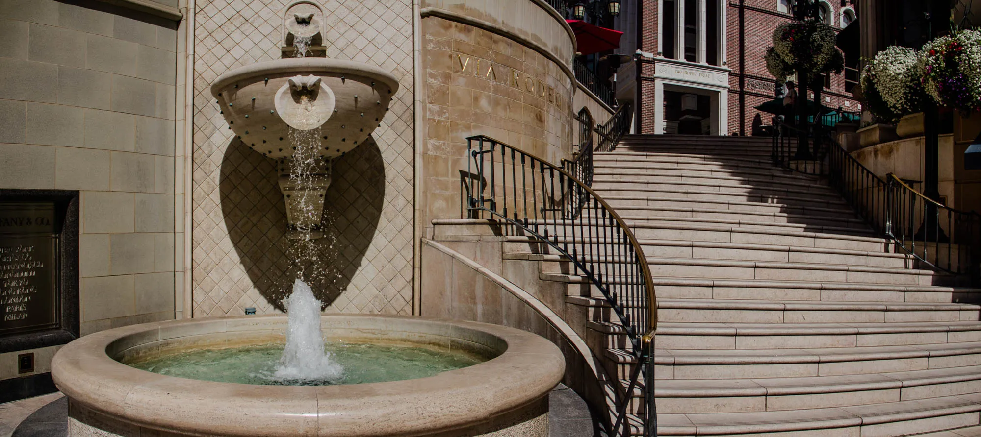 This image shows a fountain in front of a set of stairs. The fountain is made of stone and has a bowl-shaped basin. Water is flowing from a spout at the top of the fountain, creating a small spray of water. The stairs are made of stone and have a black metal railing. The railing has a curved section.  The stairs lead up to a building with the words "VIA RODEO" written on the side. There is a building to the left of the fountain, a wall with the name "TIFFANY & CO." inscribed on it.  The buildings are made of light brown stone with a diamond pattern.  There is a red awning in the background.  The image is taken from a low angle, looking up at the fountain and the stairs.