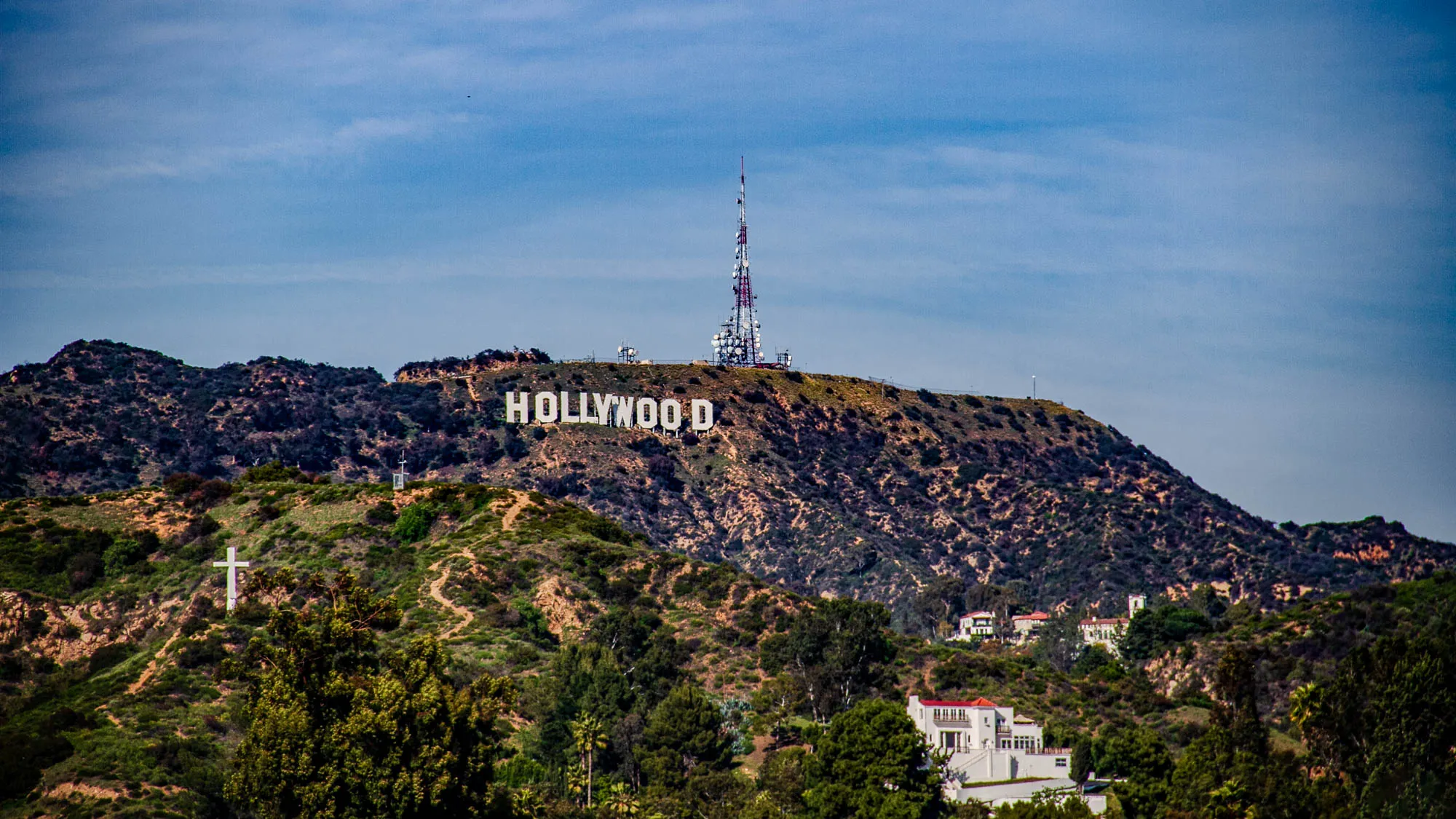 The image shows a view of the Hollywood sign on a hillside. The sign is white and stands out against the dark green bushes and trees behind it. There is a tall radio tower to the right of the sign. There are small houses on the hill in the foreground. The sky is blue with a few white clouds.  There is a small white cross on the hill below the sign. There is another white cross further down the hill on the left side of the image.