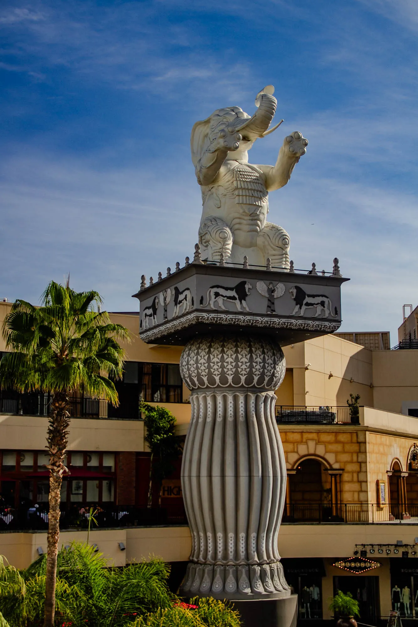 The image shows a white, elephant statue standing atop a tall, grey column in front of a building. The elephant's trunk is raised and its ears are large. The statue is very detailed, with ornate carvings and designs. The column is also intricately decorated with white and grey bands. Two lions are carved on the upper part of the column.  There are small, round light bulbs illuminating the top of the column.  In the background, a tall palm tree stands in front of a large, beige building with a balcony and decorative arches.  Behind the building, the sky is a clear, blue. There is a red sign that says "Black Swan" that is in the background of the image.