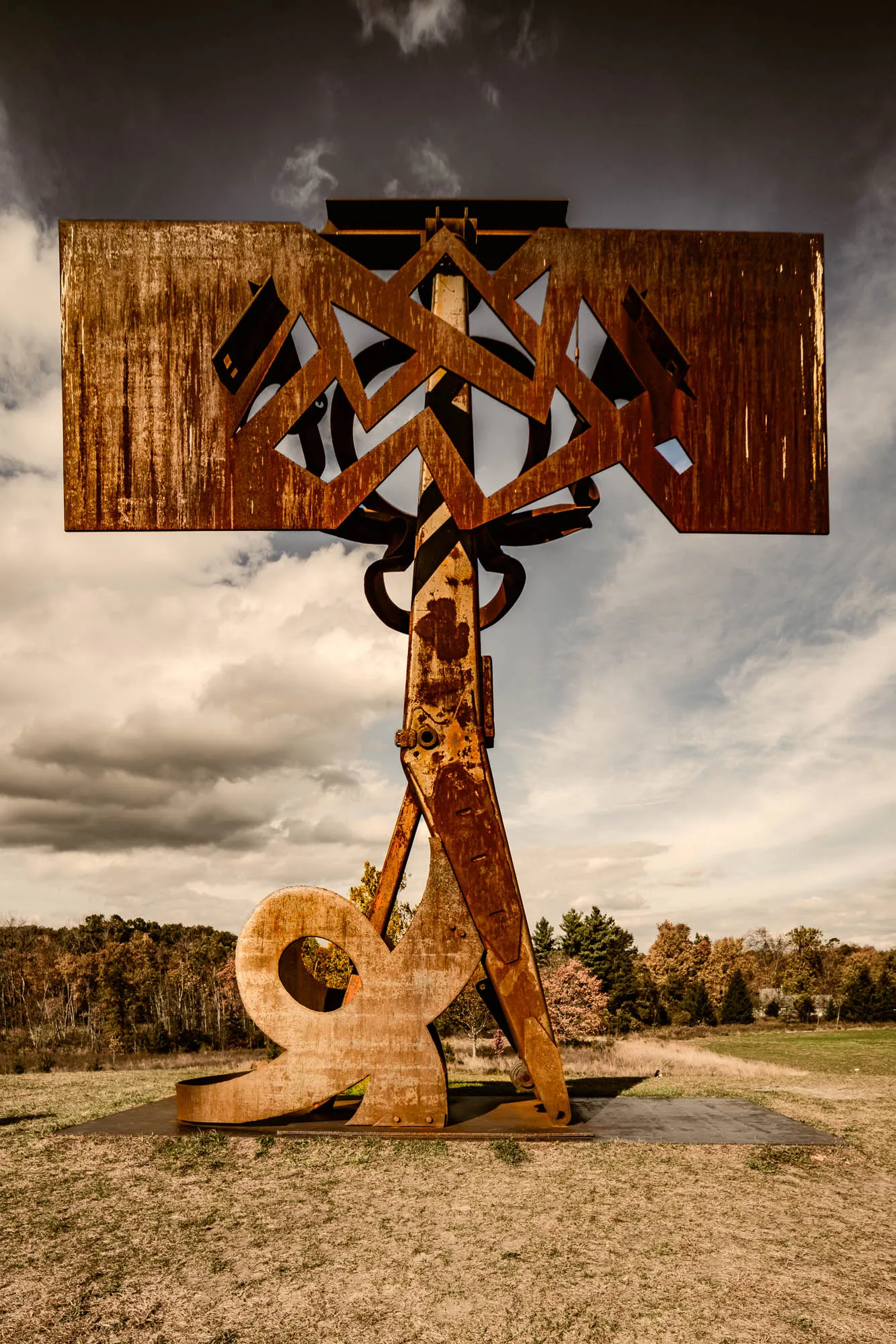 The photograph shows a large, abstract sculpture crafted from rusted metal, situated outdoors in a grassy field against a cloudy sky. The sculpture features a complex arrangement of geometric shapes and curves. At the base is a stylized shape resembling a question mark or a seated figure with a circular opening. Ascending from this base are angled metal supports that converge upwards, reminiscent of legs or stilts. These supports lead to a more intricate structure featuring circular elements and a large, rectangular metal plate at the top. The rectangular plate has a cut-out zigzag design that allows the sky to be seen through it. The rusted metal displays a rich, textured surface with variations in color, indicating weathering and age. The background is a landscape with trees in the distance, and a dramatic sky with dark clouds at the top, transitioning to lighter, cloudier areas behind the sculpture. The overall impression is one of industrial art meeting natural surroundings, with a somber and perhaps slightly apocalyptic mood.