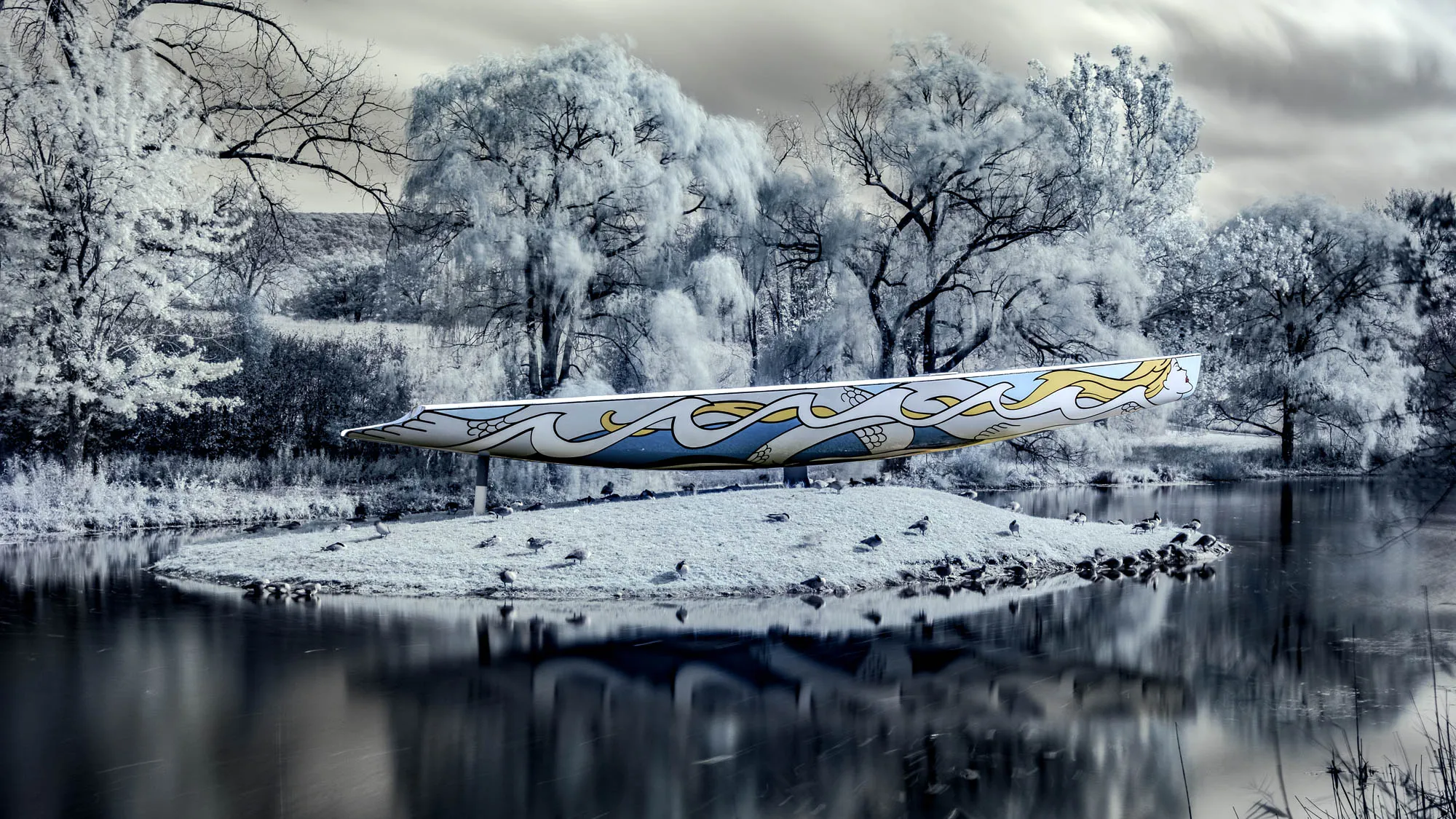 The image shows a serene waterscape in a infrared, creating an ethereal, almost otherworldly atmosphere. A sleek, decorative boat rests on a small grassy island in the center of a calm lake or pond. The boat's design features stylized waves in blue and yellow against a light background, giving it a distinct artistic flair. The island is populated by numerous birds, likely ducks or geese, some standing and others nestled near the water's edge. The water's surface is smooth, reflecting the surrounding landscape, including the island, the boat, and the silvery, frosted trees that line the banks. The trees have a textured, almost crystalline appearance, enhancing the dreamlike quality of the scene. The sky above is overcast, adding to the muted color palette and creating a soft, diffused light that bathes the entire composition.