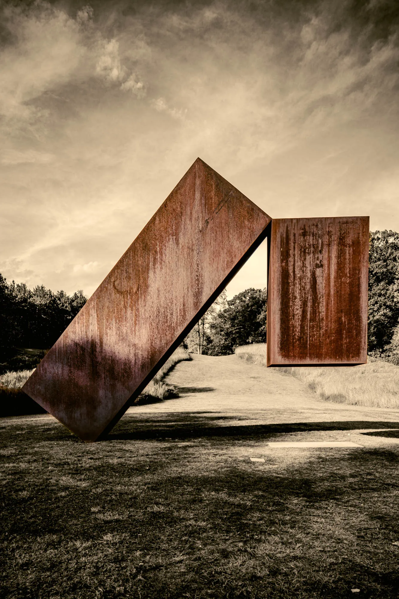 The photograph showcases a large, abstract outdoor sculpture composed of two rectangular steel plates, heavily rusted with an orange-brown hue. The scene is bathed in a sepia tone, giving it an aged or timeless feel. One rectangle leans at a diagonal, its bottom corner resting firmly on a grassy field. The other rectangle stands upright, seemingly intersecting with the leaning one, creating a sharp, angled opening. The texture of the rust is prominent, providing a sense of depth and history to the sculpture. Behind the structure, a gently sloping hill dotted with trees adds a natural backdrop. The sky, filled with soft, diffused clouds, casts subtle shadows across the landscape, adding to the three-dimensional feel of the scene. The foreground features grass of varying lengths, hinting at an open, park-like setting. The overall composition suggests a dialogue between man-made art and the natural environment.