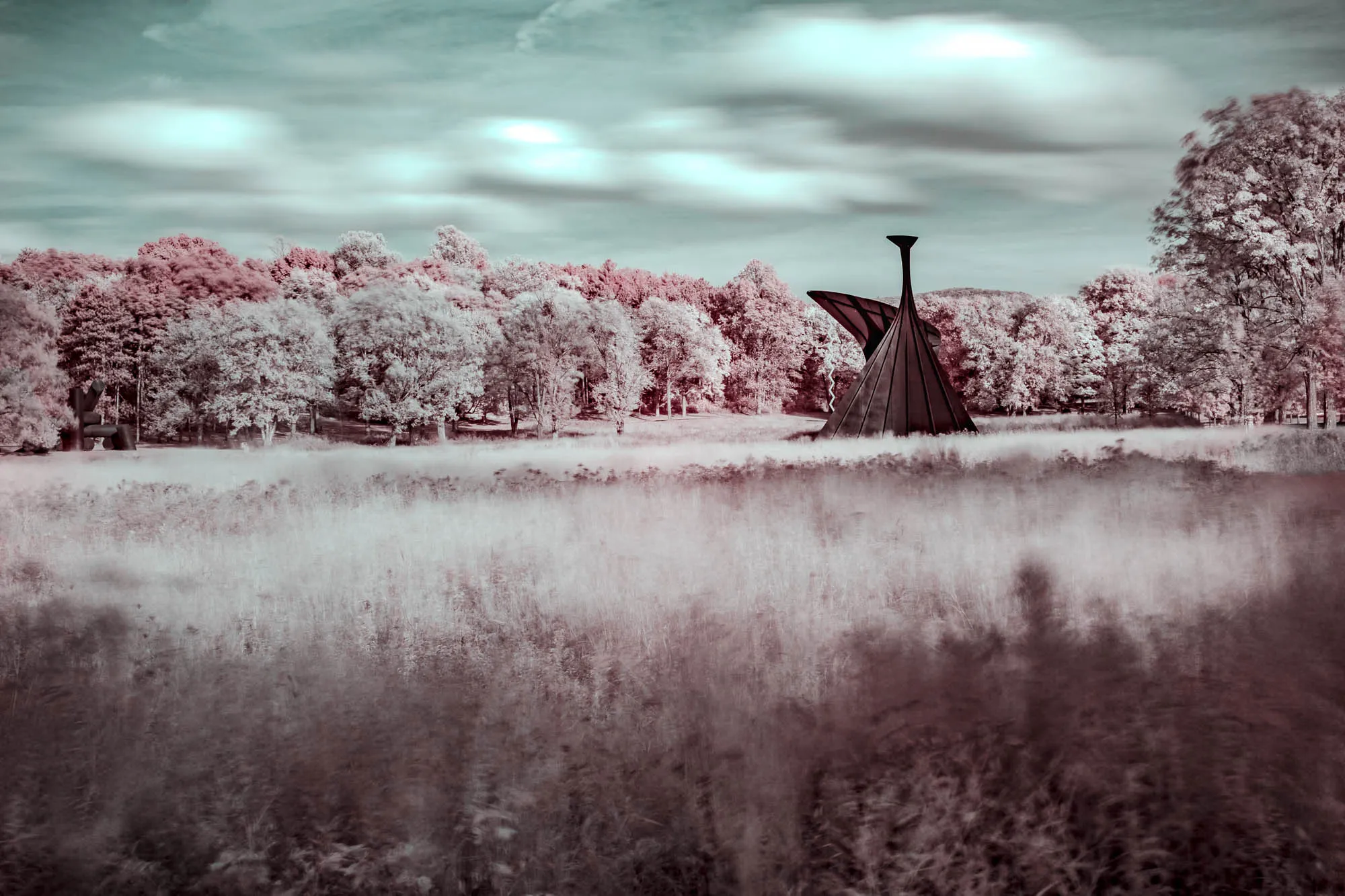 An infrared photograph of a serene landscape shows a field of tall grass in the foreground, blurring slightly with movement. Behind the grass, an abstract sculpture dominates the middle ground. The sculpture has a cone-shaped base with one large, curved wing extending upwards and outwards, topped by a slender, cylindrical element. The color palette is surreal, with the grass and trees rendered in varying shades of white and pale pink. The sky is a muted, hazy blue with soft, indistinct clouds. A line of trees forms the background, blending into the slightly blurred horizon.