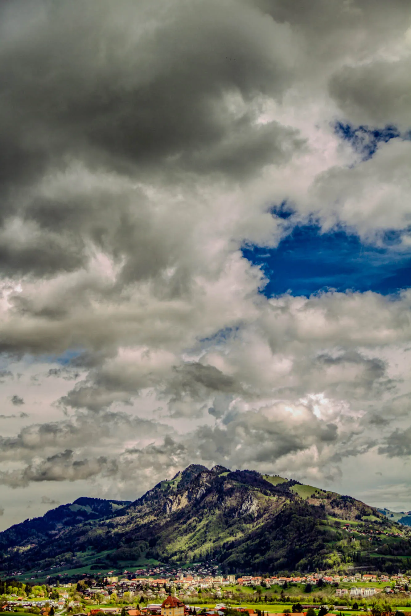 The image shows a mountainous landscape with a sky full of fluffy clouds. The clouds are a mix of white and gray, with some patches of blue sky peeking through. The mountains are covered in lush green vegetation, and there's a small town nestled at the base of the mountain.  The clouds are thick and heavy, almost as if it might rain soon.  There are trees surrounding the town and a grassy field in the foreground.