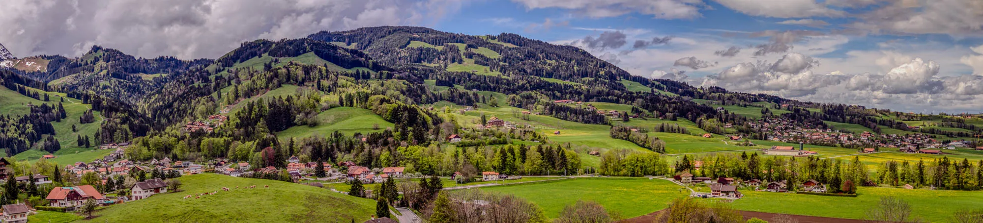 The image shows a panoramic view of a green, rolling landscape with a village nestled amongst the hills. The foreground is dominated by a lush green field with a few cows grazing. A paved road snakes through the field towards the village. The village is made up of a collection of small, mostly red-roofed houses, surrounded by trees and vegetation. In the distance, the landscape rises up into a series of hills covered in thick, green forest. The sky is a bright blue with patches of white clouds. 
