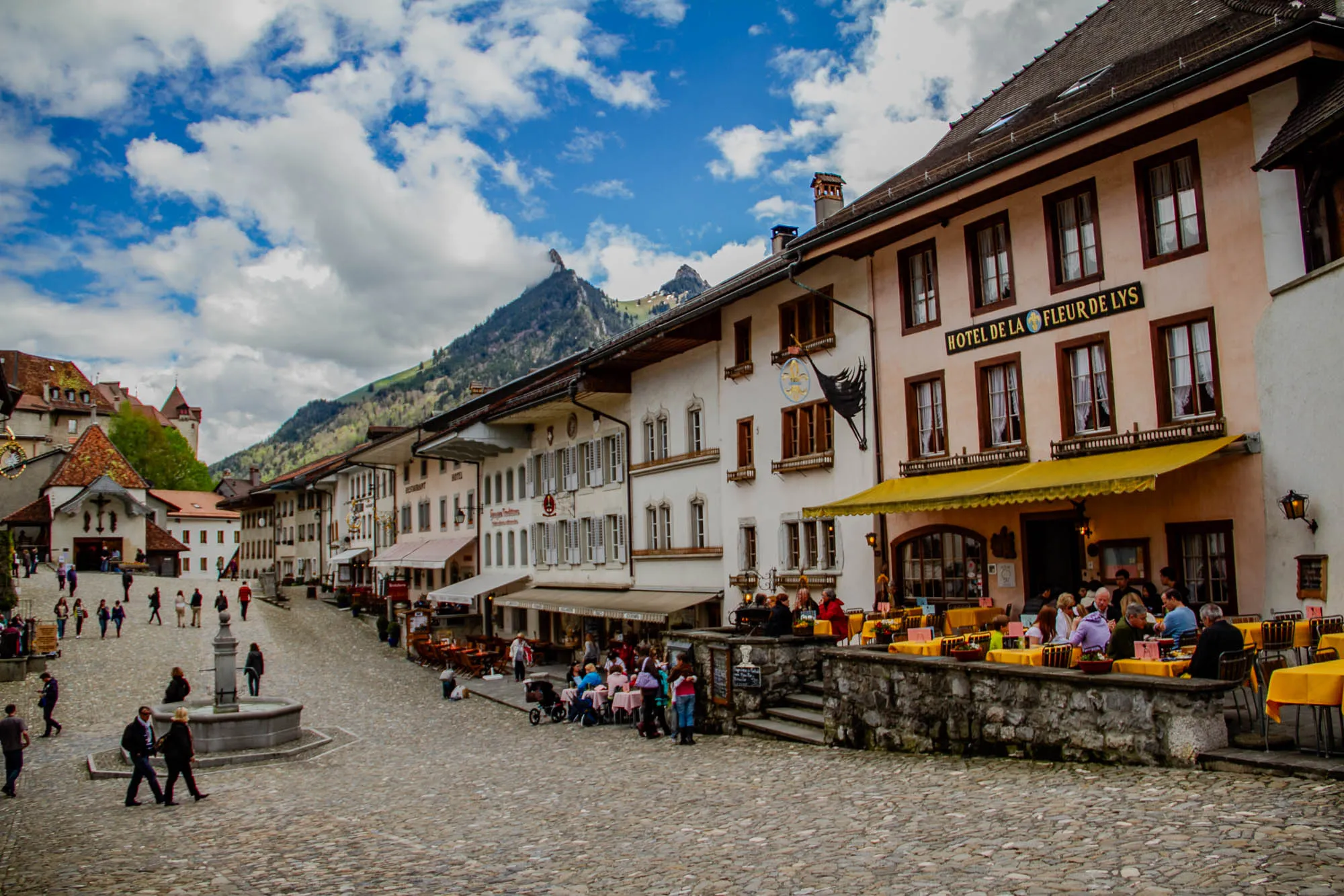 The image shows a cobblestone square in a European town, surrounded by buildings with colorful awnings and a mountainous backdrop. In the foreground, there is a fountain in the center of the square, with a few people walking around it. The square is lined with buildings, most of which have a yellow awning. The building on the right is a restaurant called "Hotel de la Fleur de Lys." The restaurant has a large sign with the name of the restaurant on it and a few people are sitting at tables outside, eating and drinking. The building has multiple windows on the second floor, all with brown window frames. In the background, there are more buildings, as well as a mountain with a clear blue sky and fluffy white clouds. The buildings are all quite old and have a traditional European style, with intricate details and colorful decorations.