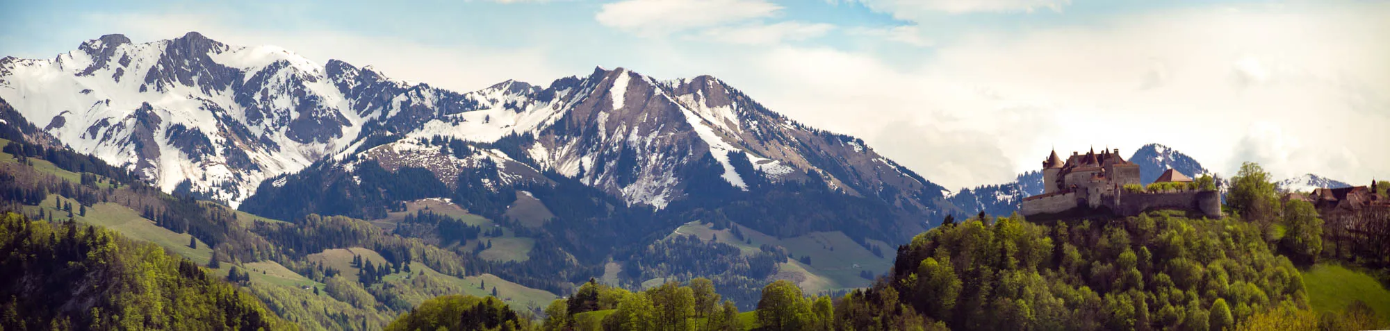 A panoramic view of a castle perched atop a forested hillside. The castle has a stone wall with towers and a pointed roof. It's set against a backdrop of snow-capped mountains. The mountains are covered in snow and have green slopes. The sky is blue with white clouds. The image is taken from a distance and shows the vastness of the landscape.