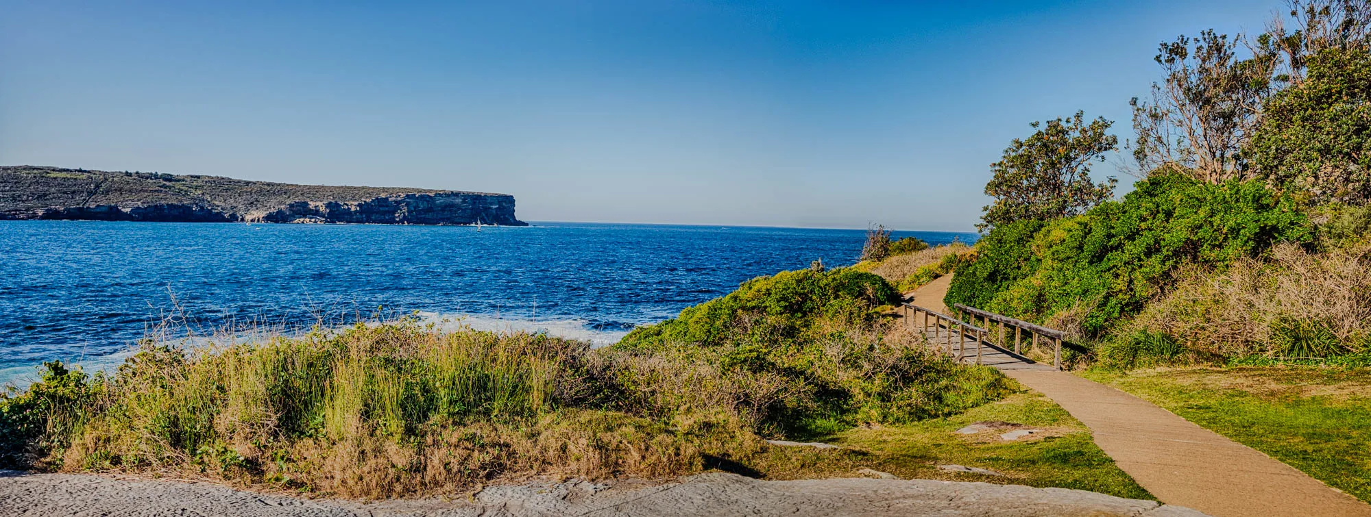 The image shows a coastal path with a wooden railing leading to a cliff edge. The path is made of light brown stones and is partially obscured by tall grass and shrubs. In the distance, a dark green cliff face sits across a vast expanse of deep blue water. The water is calm and reflective, with the sky reflecting in its surface. The sky is a clear and bright blue. There is a small boat in the distance, visible against the dark green cliff face. The scene is peaceful and serene.  