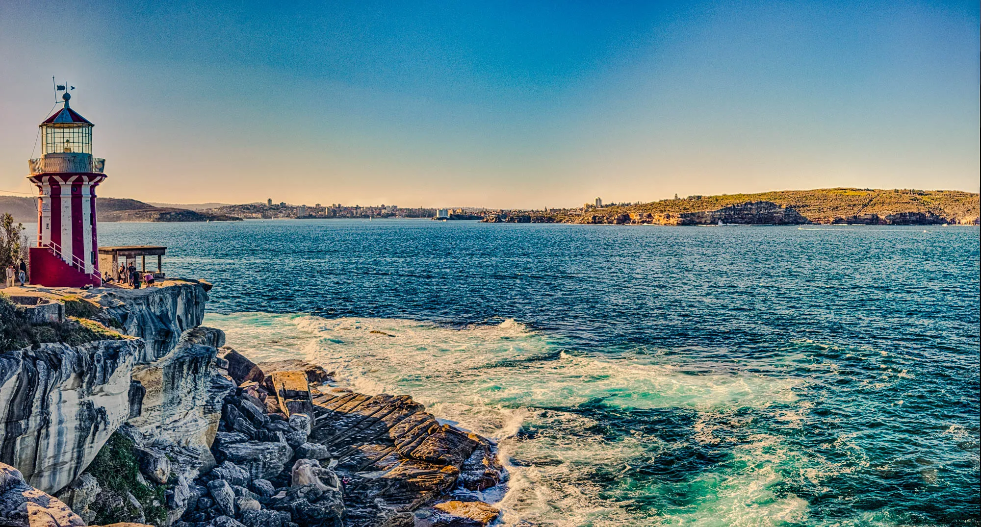 The image shows a lighthouse, standing on a rocky cliff edge overlooking a vast expanse of ocean. The lighthouse is red and white with a white top and a small black bird perched on the top. In the distance, you can see the cityscape of a coastal town across the water.  The ocean is a deep blue color, with whitecaps breaking against the rocks. The sky is clear and blue, and the sun is setting, casting a warm golden glow over the scene.  There are a few people standing on the cliff near the lighthouse, presumably enjoying the view. 
