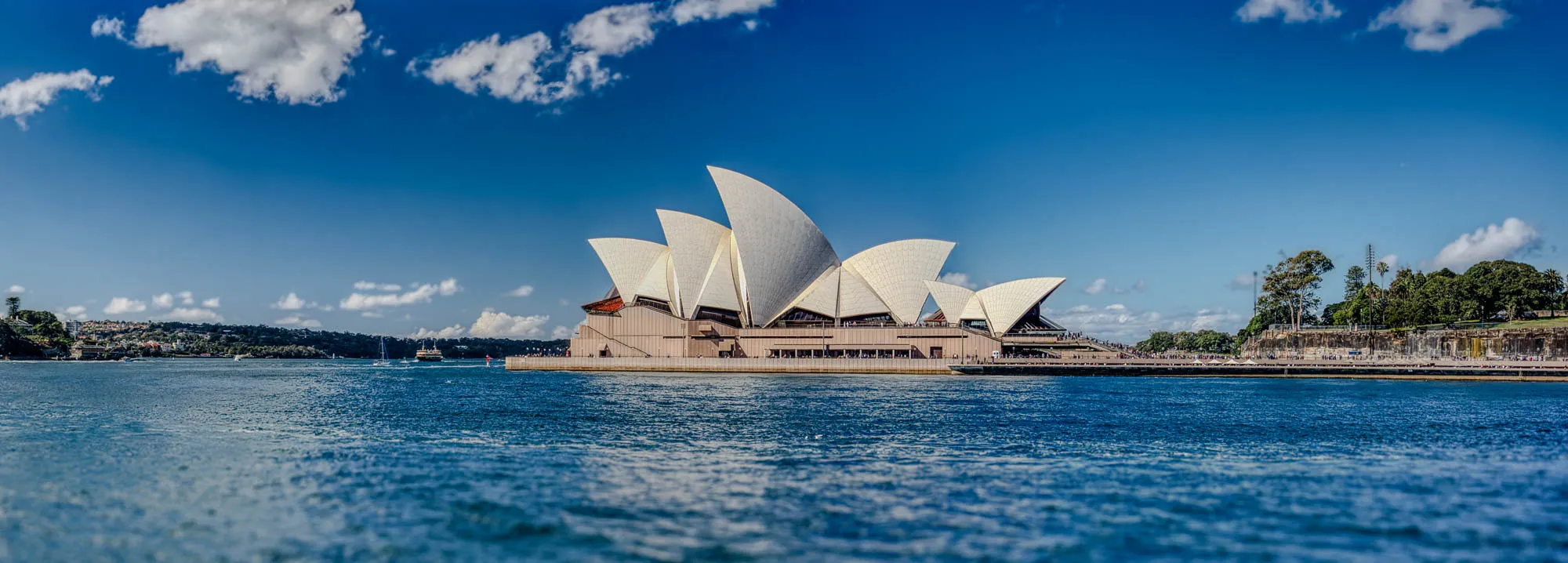 The image shows a panoramic view of the Sydney Opera House from across the water. The iconic building stands out against a bright blue sky with white fluffy clouds scattered across it. The Opera House is made up of a series of white tiled shells, each with a unique shape and curve. The building sits on a concrete platform that extends out into the water. In the foreground, the water is rippling and blue, reflecting the bright sky above. There is a small boat sailing on the water in the distance, to the left of the Opera House. In the background, there is a green forested area, with a rock cliff that slopes down to the water. The image is taken from a low angle, capturing the beauty and scale of the Opera House and its surroundings.