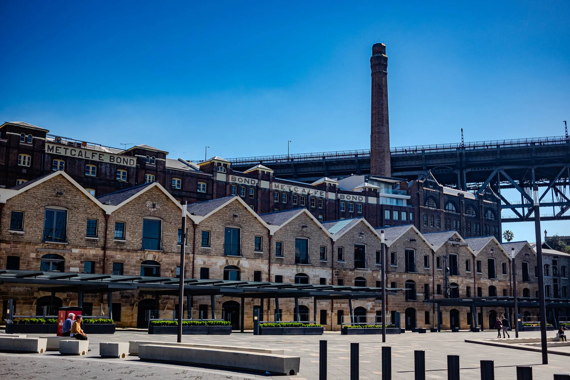 The image shows a row of brick buildings with a large metal bridge overhead and a brick chimney in the background. The buildings are made of brown brick and are lined with windows. The buildings have black roofs and grey awnings extending from the buildings in front of the windows. In front of the buildings is a wide open, paved area with grey benches. Two people can be seen walking across the paved area. The sky is a clear blue.