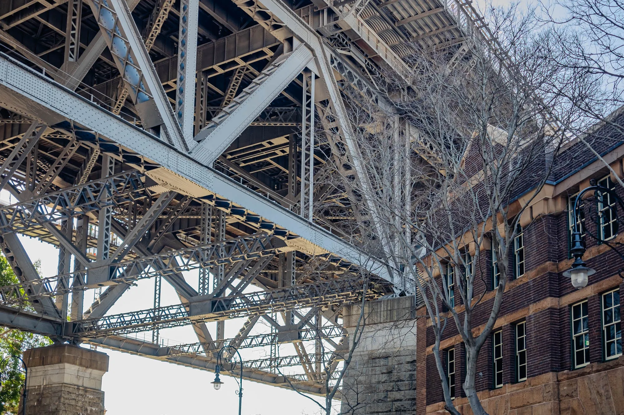 The image shows a low angle view of the underside of a large steel bridge. The bridge is made up of many intricate beams and girders, creating a complex network of triangles. The bridge is painted grey and has a light grey color scheme. The underside of the bridge is visible and you can see the many cross beams and supporting struts of the structure. The bridge is over a road or a walkway, but this is not shown in the image. Below the bridge, on the right, there is a tall brick building with many windows. There is a bare branched tree growing out of the side of the building. It is a wintery or autumnal day, as the branches are bare and the sky is clear. The building has a brown brick exterior.  A black streetlamp is attached to the building, but it is not lit. The image is well-lit with clear visibility.