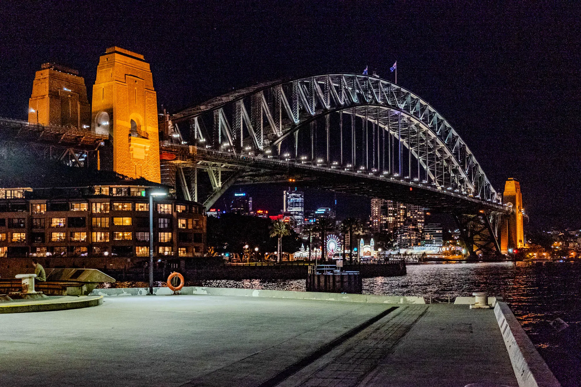 The image shows a night view of a city with a large bridge in the foreground. The bridge is made of steel and has a large arch in the middle. The bridge is illuminated with lights. The city skyline is visible in the background, and there are several buildings lit up. There is a pier extending into the water in front of the bridge. There is a person standing on the pier. In the distance, there is a Ferris wheel. The water is calm and reflecting the city lights. The sky is dark and there are some stars.