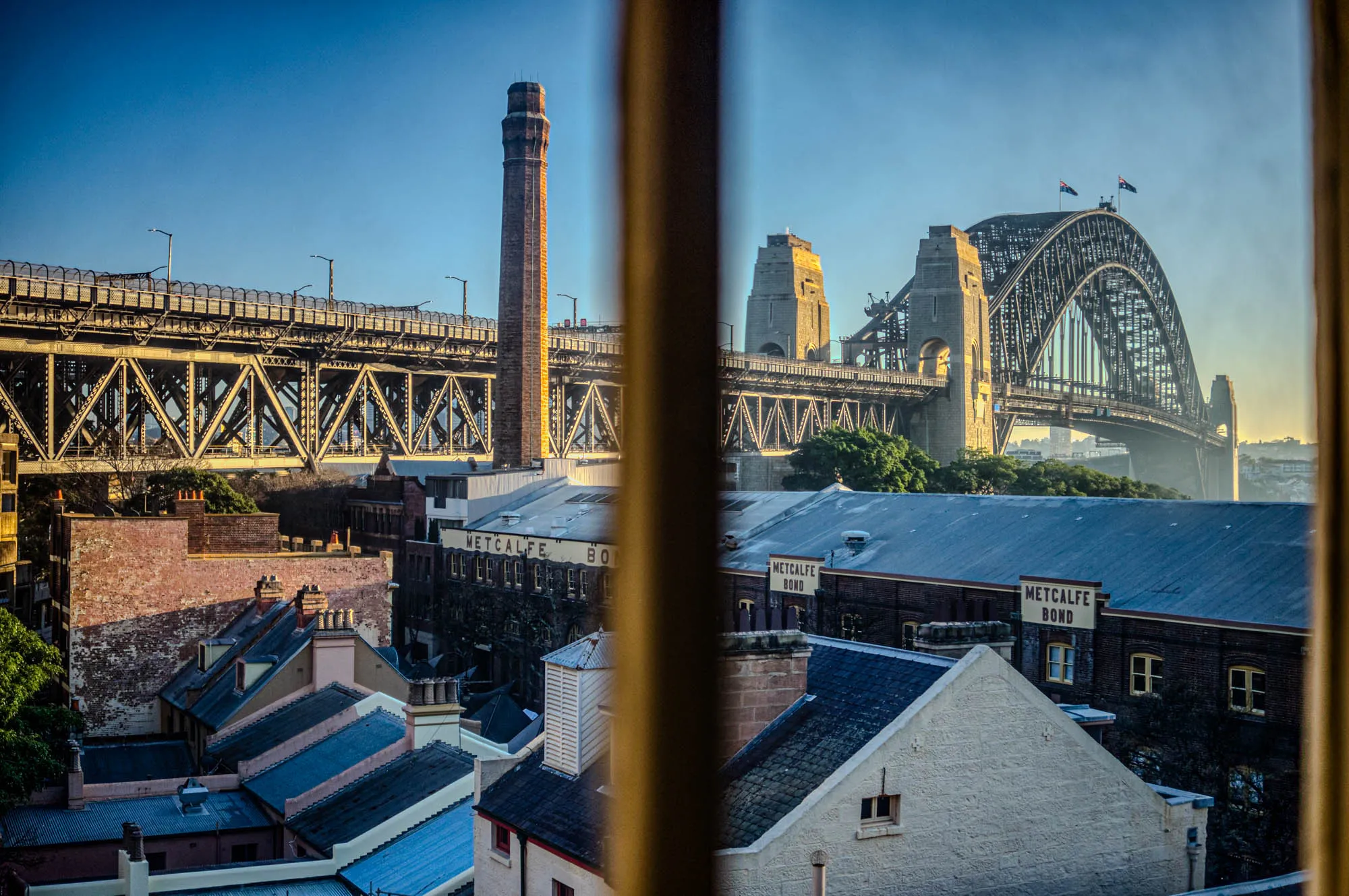 The image shows a view through a window, looking out over a cityscape. There is a large bridge in the background, which is visible through the window frame. The bridge has a single arch, and appears to be made of steel. The bridge appears to be a long way off, and is partially obscured by fog in the distance. 

In the foreground, there are a number of buildings with red brick and slate tile roofs. The buildings are all different sizes and shapes, and some have chimneys or other architectural features. The foreground buildings are closer to the viewer, and are partially obscured by the window frame. 

The image is taken on a sunny day, and the sky is a bright blue. The sun is shining brightly, and there is a slight haze in the distance. There are a few Australian flags waving on the bridge, and the name "Metcalfe Bond" is visible on a building in the foreground.  