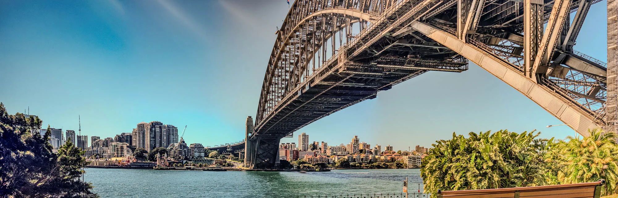 The image shows a low angle view looking up at the underside of a large steel arch bridge. The bridge spans a body of water with a city skyline in the distance. You can see the complex steel framework of the bridge, including the various beams, girders, and supports. The city skyline is visible through the bridge, with several tall buildings and other structures. The water below the bridge is calm and appears to be reflecting the sky. The foreground of the image shows some lush green trees and foliage. The sky is a clear blue with a few white clouds. This is a beautiful image that captures the grandeur of this iconic bridge and its surrounding cityscape. 
