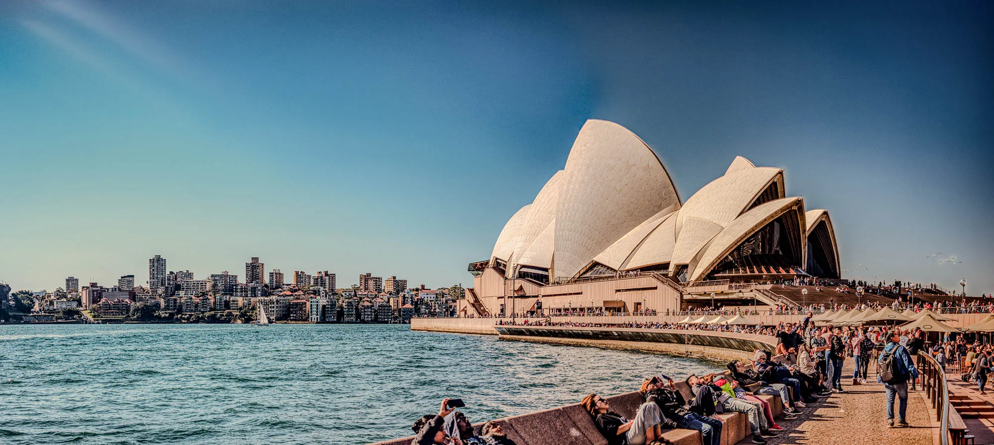 The image shows the Sydney Opera House, a famous landmark in Sydney, Australia. It is a large building with a distinctive design of multiple white, sail-like shells. In the foreground, there are many people sitting on a stone path in front of the Opera House.  The path extends along the waterfront, and in the background, a cityscape of buildings and a calm, blue body of water can be seen. The sky is a clear, light blue, and the sun is shining brightly.  There are a few white sailboats visible in the distance on the water.