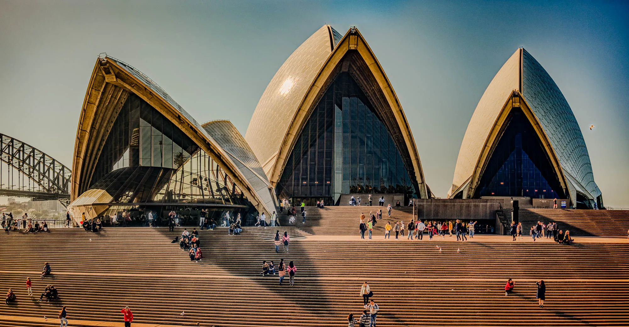 The image shows the Sydney Opera House. The building is a series of white, sail-like structures made of tile and is in the shape of a series of shells. The Opera House sits at the end of a wide, paved staircase. There are many people walking up and down the stairs and some sitting on the steps, enjoying the view of the building. The sun is shining and the sky is blue. Behind the Opera House is a metal bridge. There are many people on the bridge and in the surrounding area.