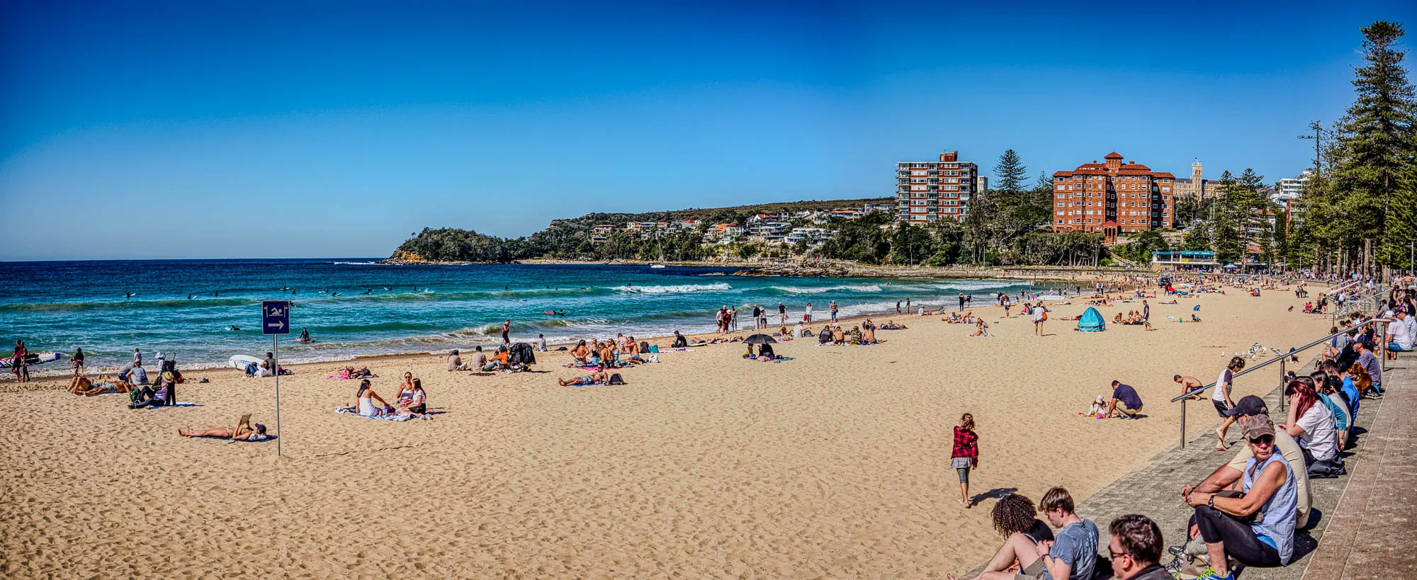 The image is a wide shot of a beach on a sunny day. The beach is packed with people enjoying the sun. Many people are in the water, some are laying out on the sand, others are swimming, and some are surfing. The water is clear blue and the sand is light brown and mostly empty, but there are some colorful umbrellas and towels scattered on the sand.  There are tall trees along the far side of the beach and buildings in the distance.  The sky is blue and cloudless. There is a blue sign with an arrow pointing to the right which is likely a sign for the beach. The beach is very busy. There are people of all ages and some are sitting on the edge of the beach, possibly on a concrete wall, looking out at the ocean.