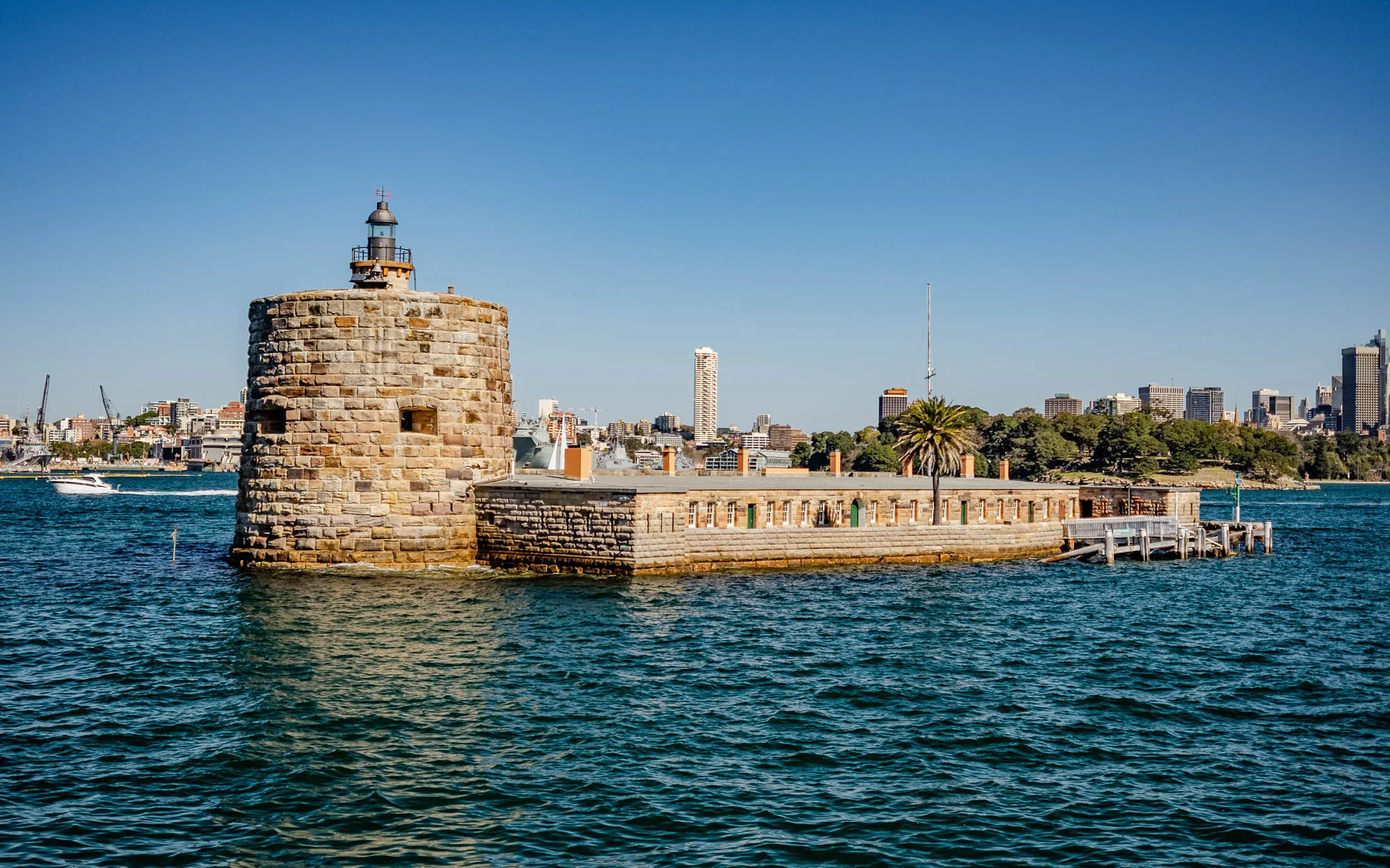 The image shows a stone fort with a lighthouse on top of a tower. The fort is built on an island with a small dock and a wooden walkway leading to it. The water surrounding the fort is dark blue and has ripples on the surface. In the background, there is a city skyline with buildings of various heights and a clear blue sky. The fort appears to be old and historic, with weathered stone walls and a tower with a lighthouse on top. The image evokes a sense of history and maritime tradition.  