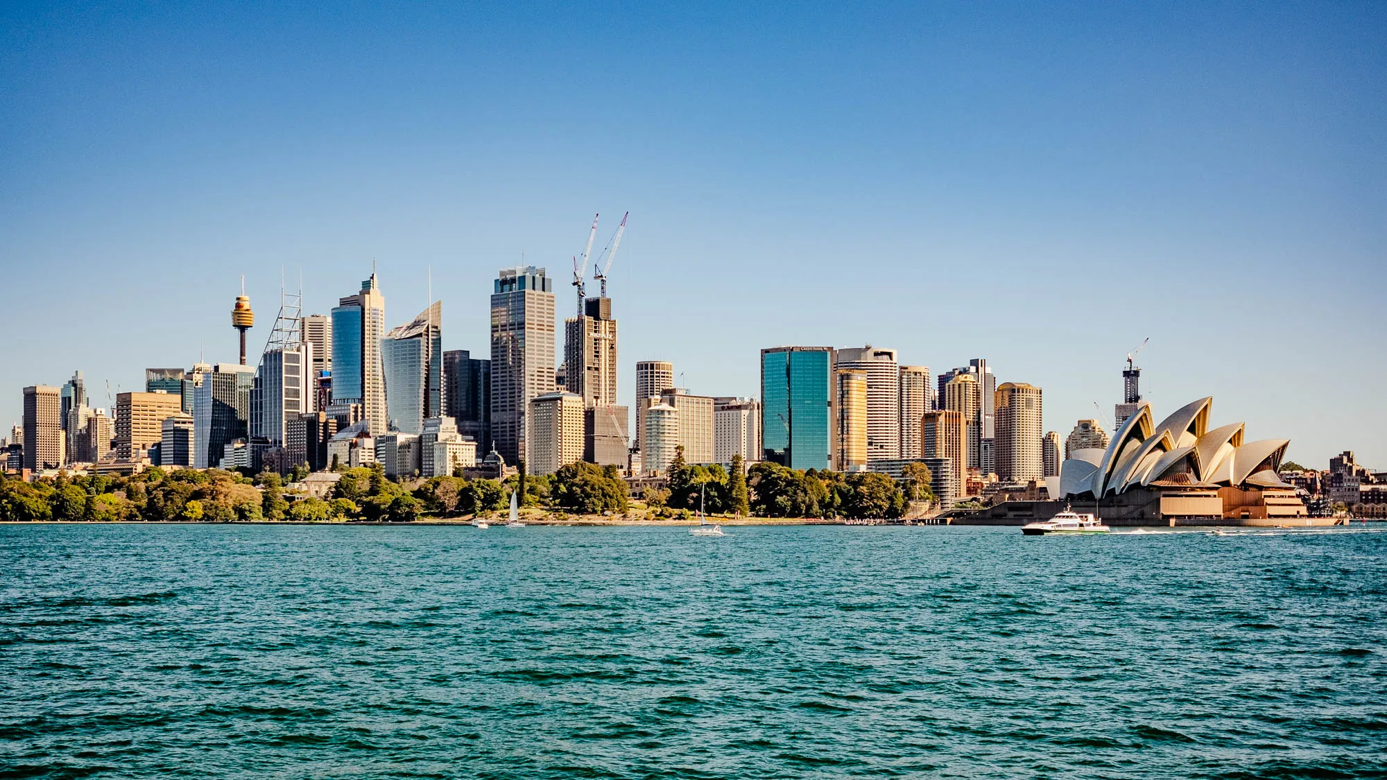 The image shows a panoramic view of Sydney, Australia. In the foreground, there is a large body of water. In the background, the city skyline is visible. The skyline includes tall buildings, with the Sydney Opera House in the right-most part of the skyline.  A few sailboats and a ferry are visible on the water in the foreground. The sky is clear and blue.