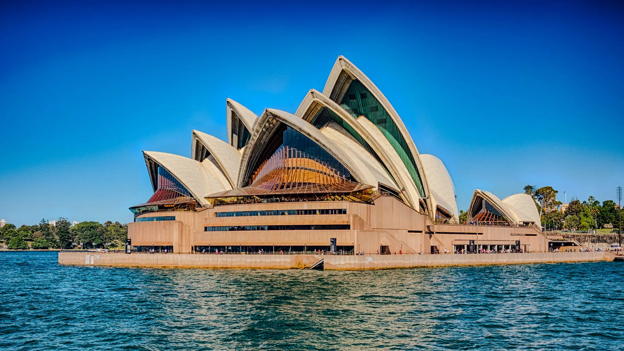 The Sydney Opera House is a large building with a unique design featuring a series of white, curved shells. The building is constructed of concrete and features a large number of windows. The structure is situated on a waterfront with a blue body of water.  A small number of people are visible near the water's edge. The sky is clear blue.  