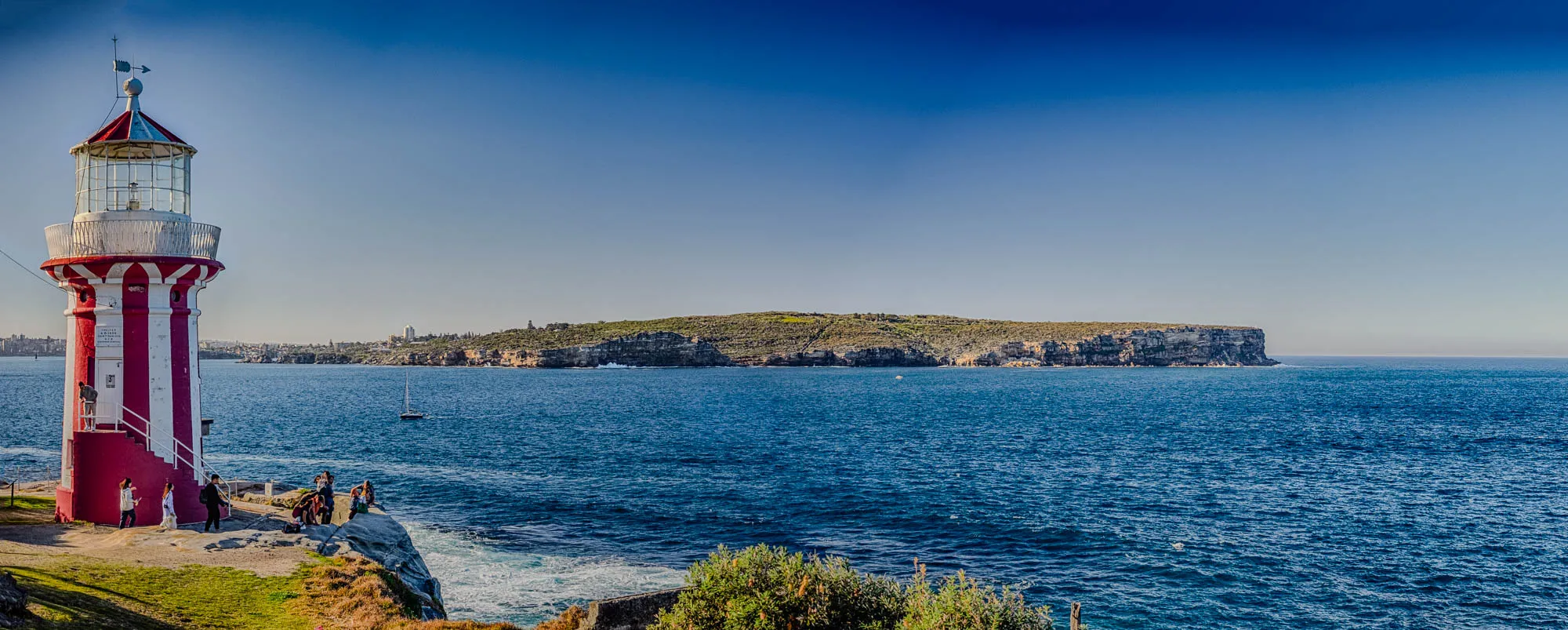 The image shows a red and white lighthouse with a glass lantern on top. The lighthouse is located on a cliff overlooking a body of water. There are a few people standing near the lighthouse, and a sailboat is in the distance. The sky is a bright blue, and there is a small island on the horizon. In the foreground, there are some green plants growing. The lighthouse is on the left side of the image.