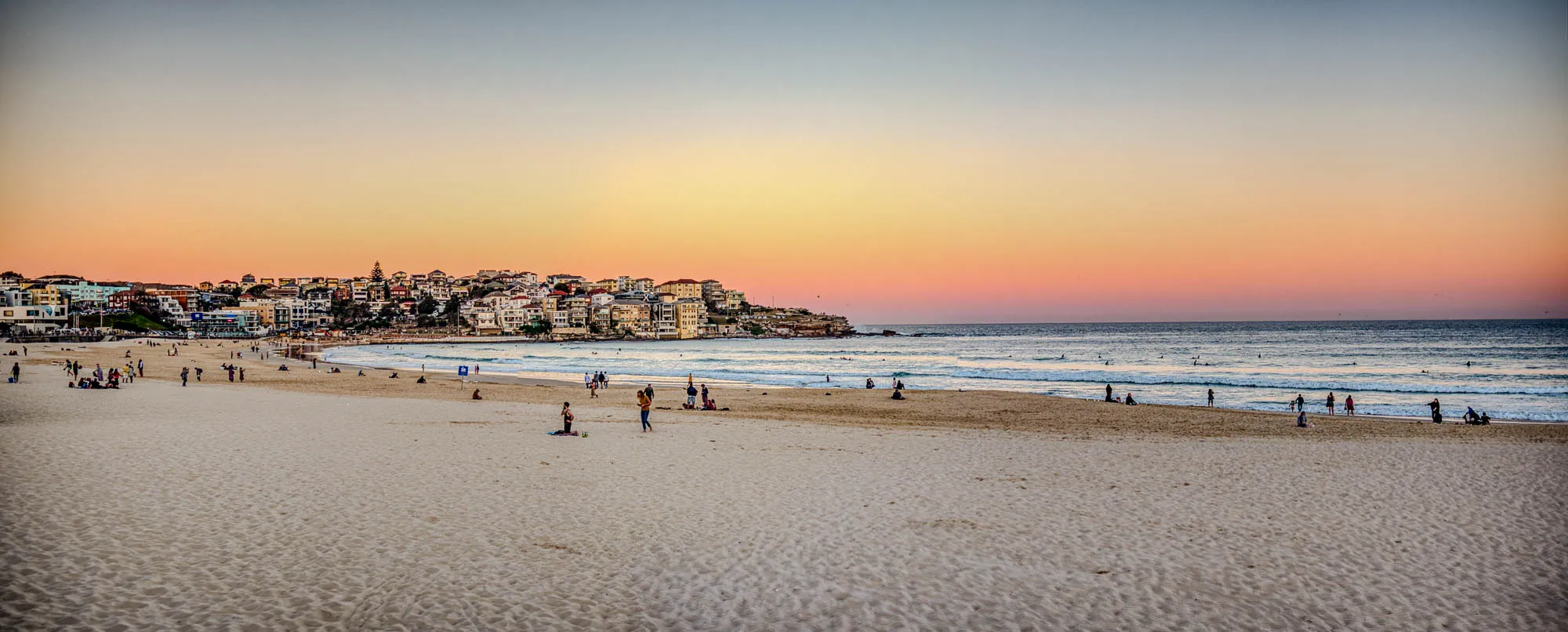 The image shows a beautiful beach scene at sunset. The sun is setting over the horizon, casting a warm glow over the sky. In the distance, a city skyline can be seen with many buildings and houses. The beach is mostly empty, with a few people scattered about.  Some people are sitting on the sand and others are walking along the shore. The ocean is a calm blue, and the waves are gently rolling in. The scene is peaceful and serene, with a sense of relaxation and tranquility. The overall mood of the image is one of beauty and wonder.