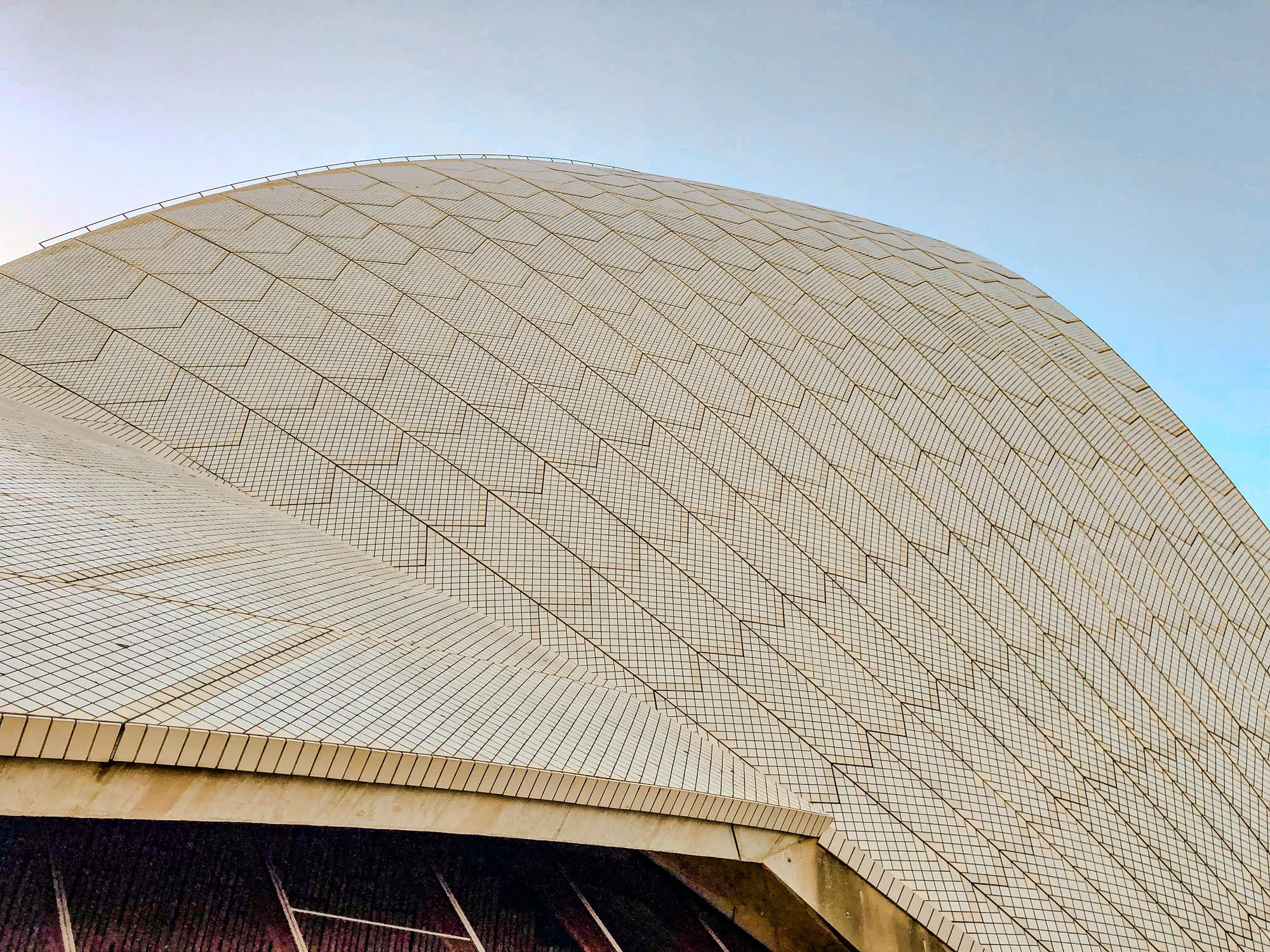 The image is a close-up of a curved, tiled surface. The tiles are white and arranged in a grid pattern, creating a sense of depth and texture. There are thin black lines running diagonally across the surface, forming a larger pattern of overlapping squares. The surface curves upwards, creating a sense of  monumentality. The background is a clear blue sky.  The surface might be a section of the Sydney Opera House.