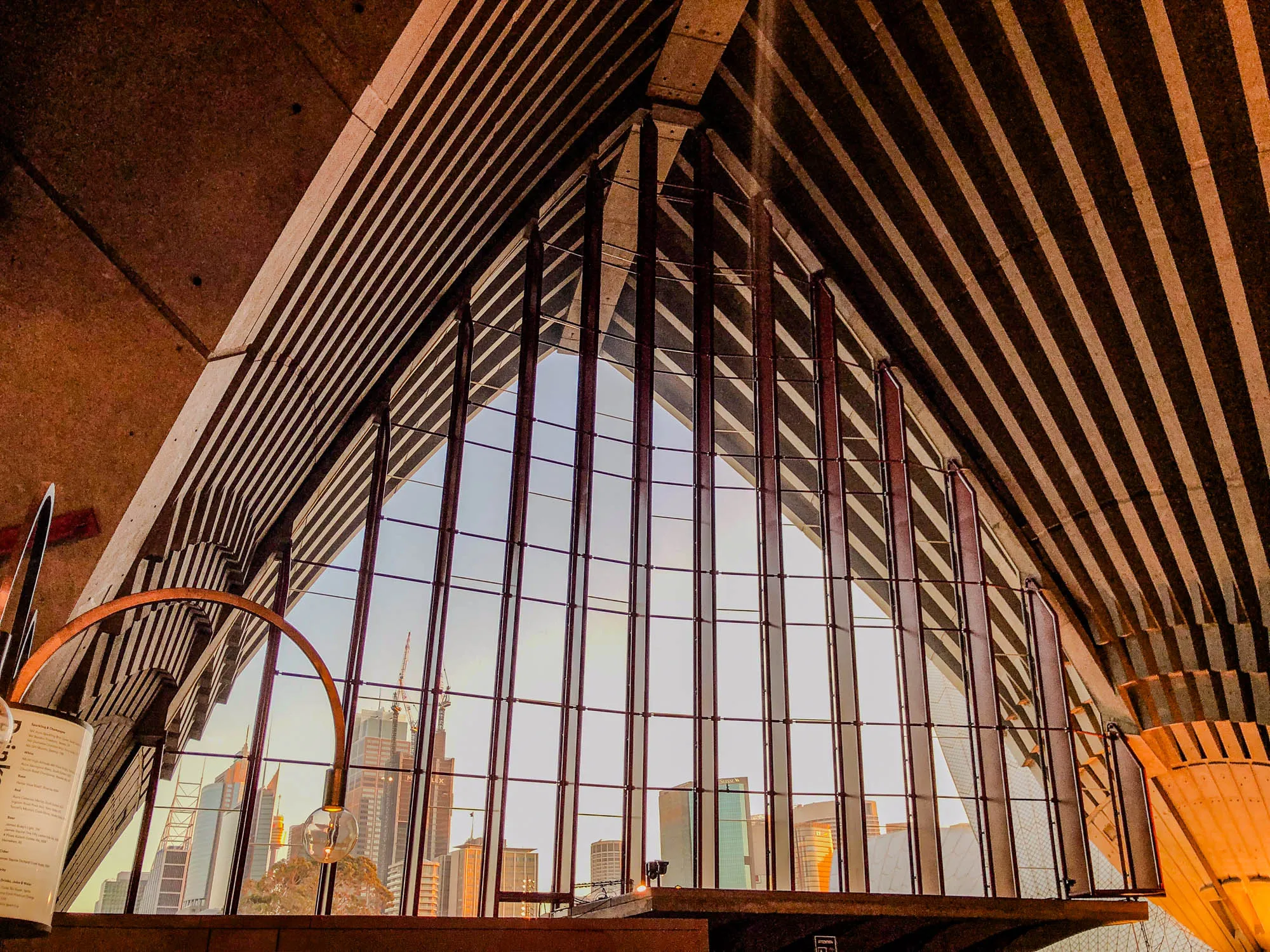 The image is of a large window in a building with a view of a city skyline. The window is made up of a grid of vertical and horizontal bars. The city skyline is visible through the window, with a few tall buildings and several shorter buildings visible. The sky is a pale blue color, and the sun is setting in the background.  The roof above the window is composed of a pattern of horizontal stripes and diagonal lines. It is made of a wood-like material.  A large metal arc is suspended from the roof near the bottom of the image and extends into the window.