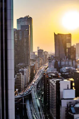 A tilt-shift timelapse of the sunset in Tokyo.  The sun is shining brightly in the sky, which appears golden yellow.  In the foreground is an elevated highway with cars driving on it.  To the left, a tall building dominates the scene. In the distance, there are more buildings. It appears to be a sunny day. The sky is bright blue. The photo was taken at a wide angle. It is a horizontal photo.  The image is in focus and clear.  The colors are natural.