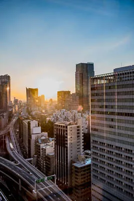 An aerial view of a city at dusk. In the center of the image is a multi-lane highway stretching from the foreground to the background. The highway is lined with tall buildings on either side. The buildings are all gray or brown and many have windows. There is a lot of traffic on the highway, visible as blurry streaks of light. The sky is a light blue, indicating dusk. In the background, the buildings are more blurry and indistinct. The photo was likely taken from a tall building or helicopter.