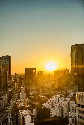 A timelpase of the sunset behind Mt. Fuji with Tokyo in the foreground. The sun is setting in the distance, casting a golden glow over the buildings. There are many tall buildings in the image, including a large skyscraper in the right background. There are also many smaller buildings in the foreground. The scene is busy and full of life.  A highway runs through the city, with cars driving on it. The scene is tranquil and peaceful.  The sky is a soft blue, with a few clouds visible.  The buildings are mostly made of glass and steel, with some older brick buildings mixed in. There are trees and plants scattered throughout the city. The overall effect is one of beauty and grandeur. 
