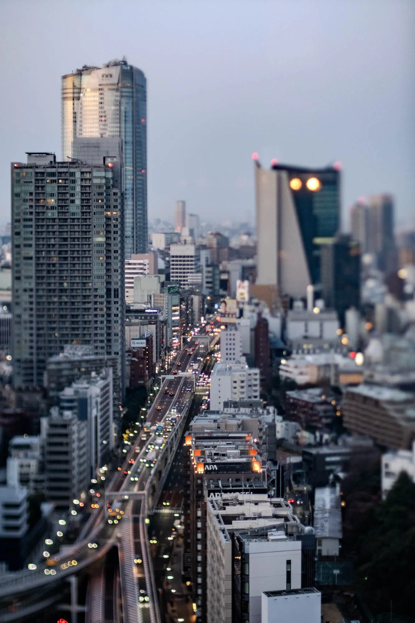 The image is a cityscape with a street full of cars. The street is lined with tall buildings, and the sun is setting in the background. On the left side of the street, you can see a building with large, reflective windows. The windows are arranged in a grid pattern, and they reflect the sun, causing the building to appear very bright. In the middle of the image, a tall building with a sign that reads "TIBS" is partially visible through the other buildings in the background. The street is busy with traffic, including cars, trucks, and vans. There are people walking on the sidewalks, and the street is lined with lampposts. The image has a warm, golden glow, and it is a beautiful representation of a bustling city.