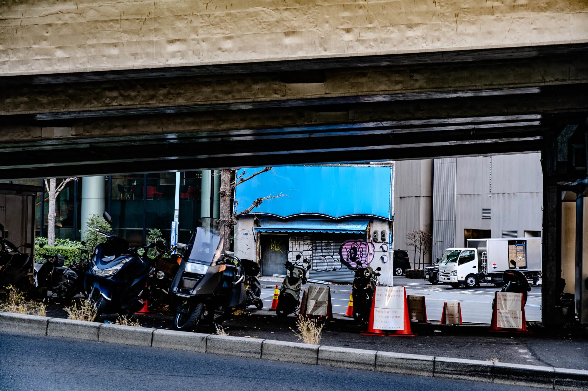 The image shows a view from underneath a concrete overpass. Looking straight ahead is a paved road with a concrete curb on the left. There are five motorcycles parked along the curb on the left side of the road. They're mostly black, and some have large windshields. The street is lined with orange construction cones and signs with Japanese writing. The road ends at a busy street where a white delivery truck is parked on the right side. The delivery truck has a large blue Pepsi logo on the side. The delivery truck is parked in front of a large building. The building is made of light-colored concrete with large windows. There is a large blue billboard on the top of the building with some graffiti below it. There is a small tree growing in front of the building. There is an overpass in the foreground of the image. The overpass is made of dark concrete and has a rough texture.