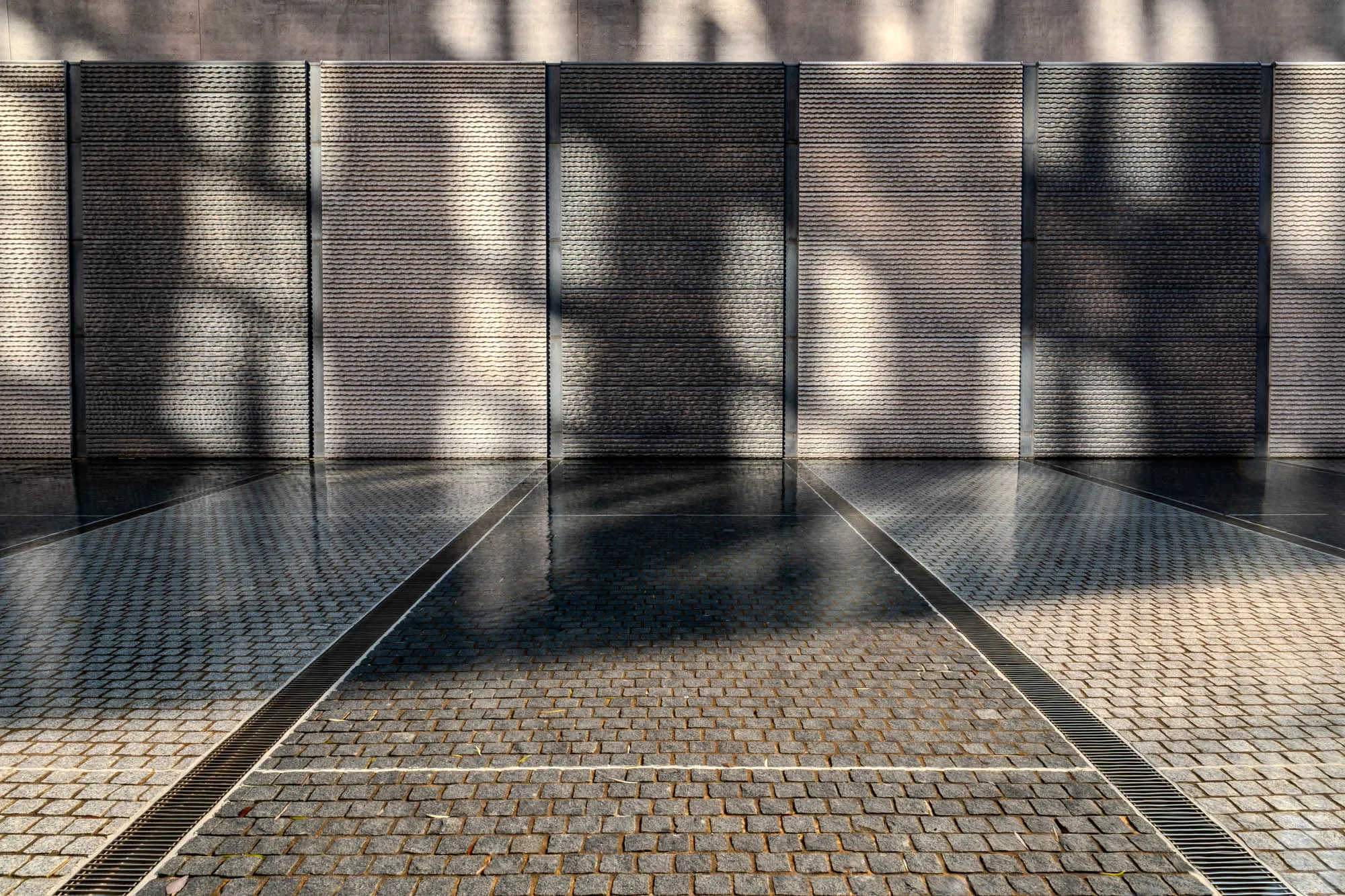 The image shows a courtyard with a wall in the background. The wall is made of three sections of gray stone with thin black lines separating each section. The wall is textured and has a pattern of horizontal grooves running across the surface. The texture of the stone is not uniform, and there are dark spots interspersed throughout the lighter gray of the stone. The wall is reflecting the light of the sun, which is casting shadows on the wall. There is a small shallow pool of water in the foreground. The water reflects the shadows cast by the wall, and is very still, so the reflection is almost a mirror image. In front of the pool is a cobblestone walkway made of squares of light gray stone with a darker gray between the squares.  There are two metal drainage grates to the left and right of the walkway. The grates have a pattern of parallel lines and are made of stainless steel.