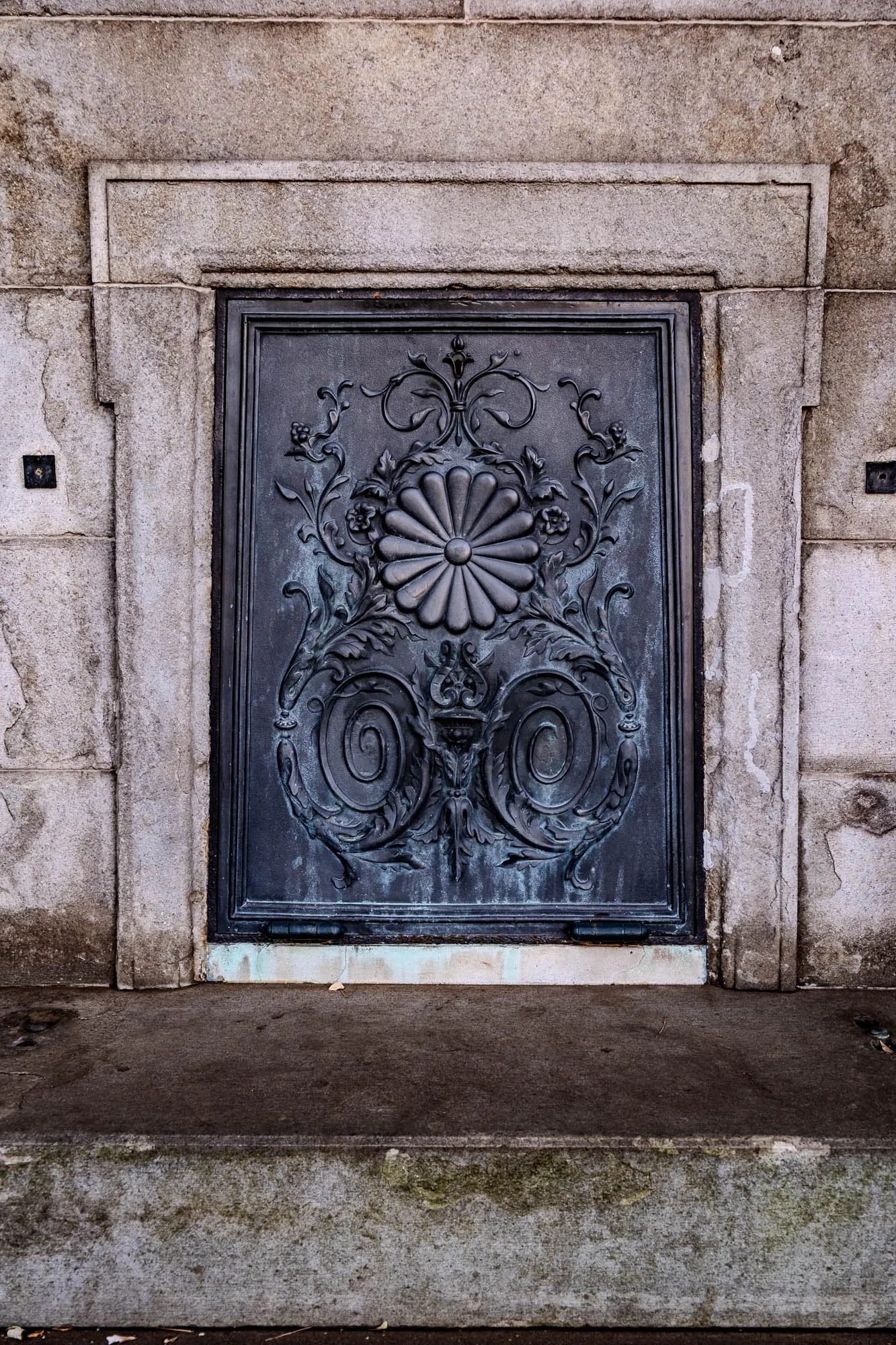 The image shows a dark metal door with a detailed design on it. The door is set within a stone frame with a large step in front of it. The metal design is embossed and features a large floral motif that resembles a chrysanthemum or similar flower. It is centered and surrounded by swirling vines. The door has a smooth surface with a hint of a teal patina. The frame and the step have a rough texture and are made of a light gray stone, similar to concrete, with some weathering. The step is covered with dirt and moss. There is a small dark rectangle on the left side of the frame, near the top, which may be a door handle. The door appears to be closed and is situated in a wall that is also made of a similar light gray stone.