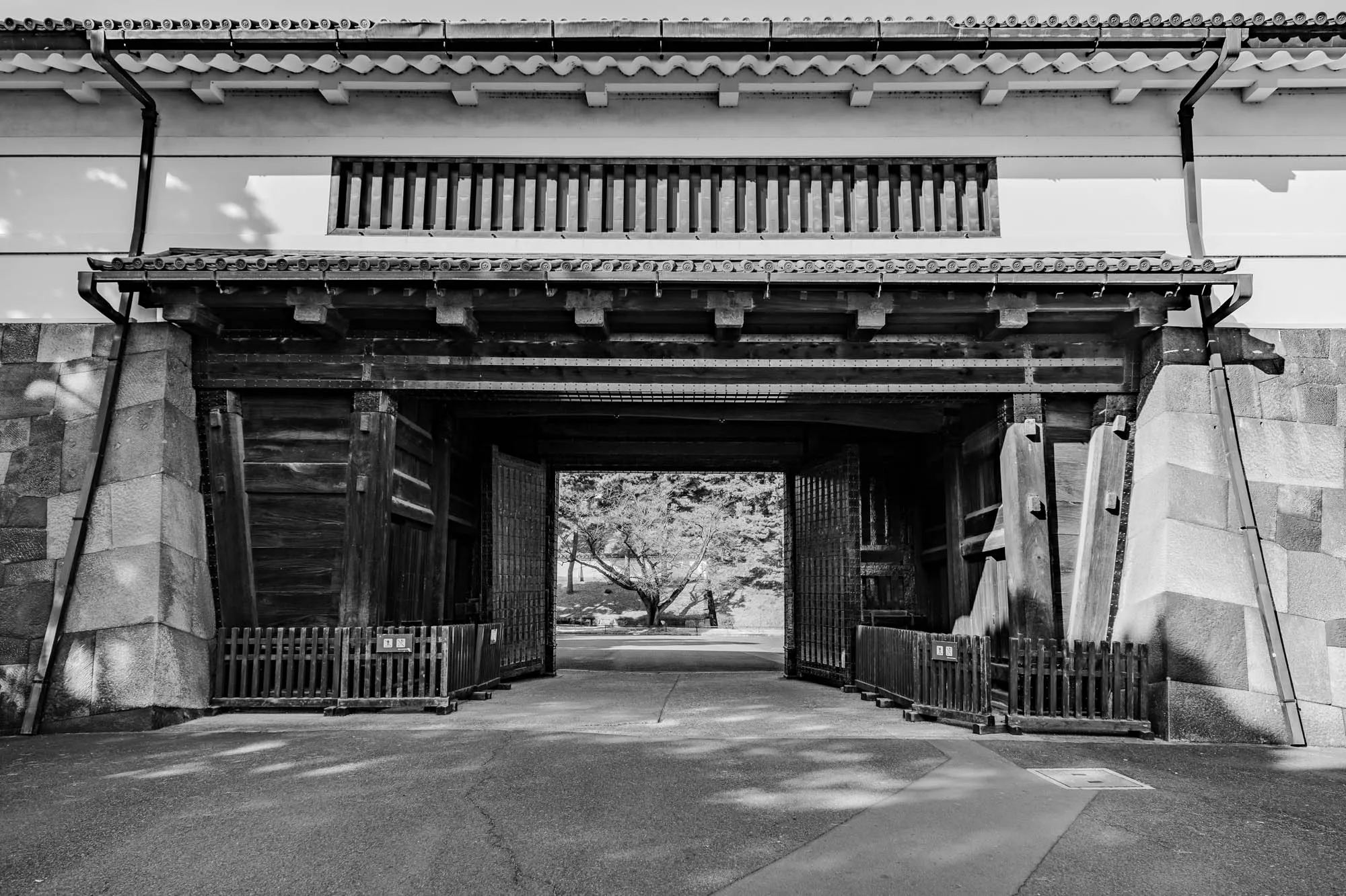 The image is a black and white photograph of a large wooden gate with two sets of doors, in a stone and wood structure. The gate is open, revealing a view of a tree in the distance. The gate has a simple, traditional Japanese design and is made of dark wood. It is situated on a walkway made of stone. The gate is set within a stone and wood building, with a white roof and a black gutter running down the side. The building appears to be made of small stone blocks, laid in a pattern. There are two small fences on either side of the gate. The photo appears to be taken on a sunny day, as the shadows cast by the structure are quite sharp.