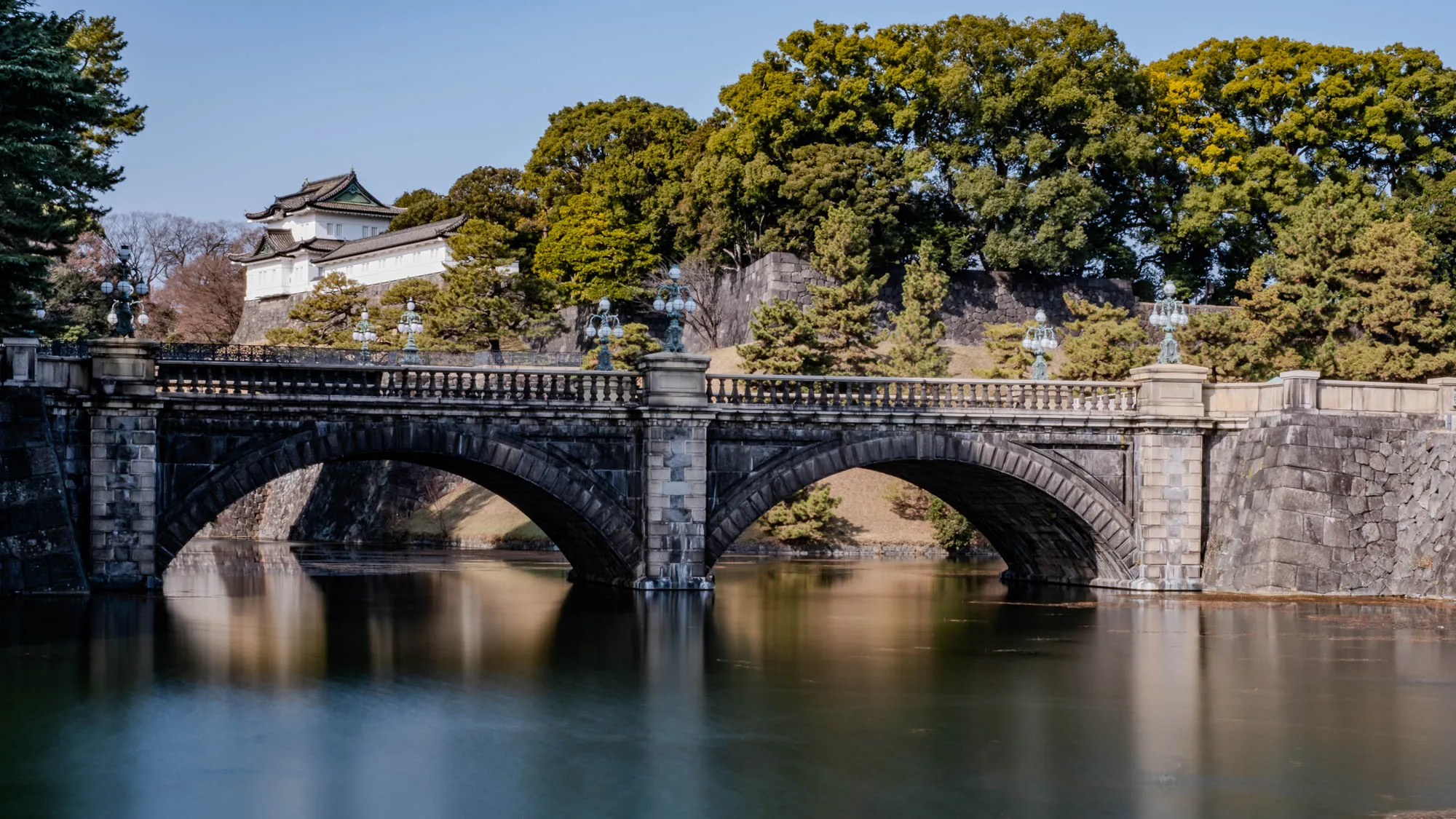 The image shows a stone bridge with two arches, spanning a calm body of water. The bridge appears to be made of gray stone with a smooth, slightly weathered surface. The arches of the bridge are tall and wide, and the bridge itself is relatively narrow. On either side of the bridge are stone walls, possibly part of a larger structure or fortification. There are trees in the background, suggesting a park or garden setting.  The sky is a clear blue, indicating a sunny day. The water is still and reflective, making it difficult to see details of the bottom. There are also a few lampposts on the bridge, adding a decorative element to the scene. The overall impression is one of peace and serenity.  The bridge seems to connect two sections of a larger complex, and the setting is likely a historical or cultural site. 
